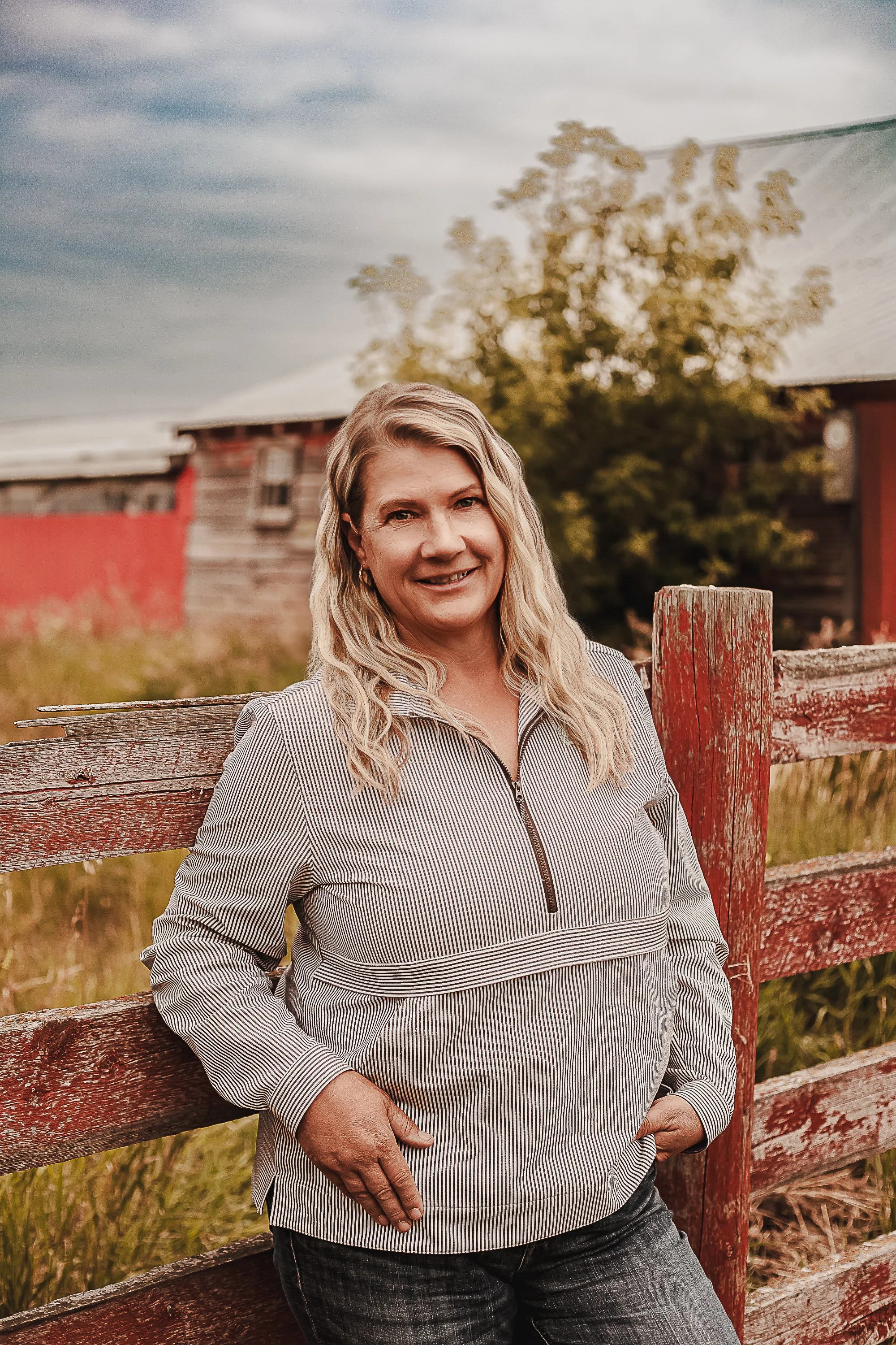A woman standing outdoors near a red wooden fence, with a barn and a tree in the background, during daytime with cloudy sky.
