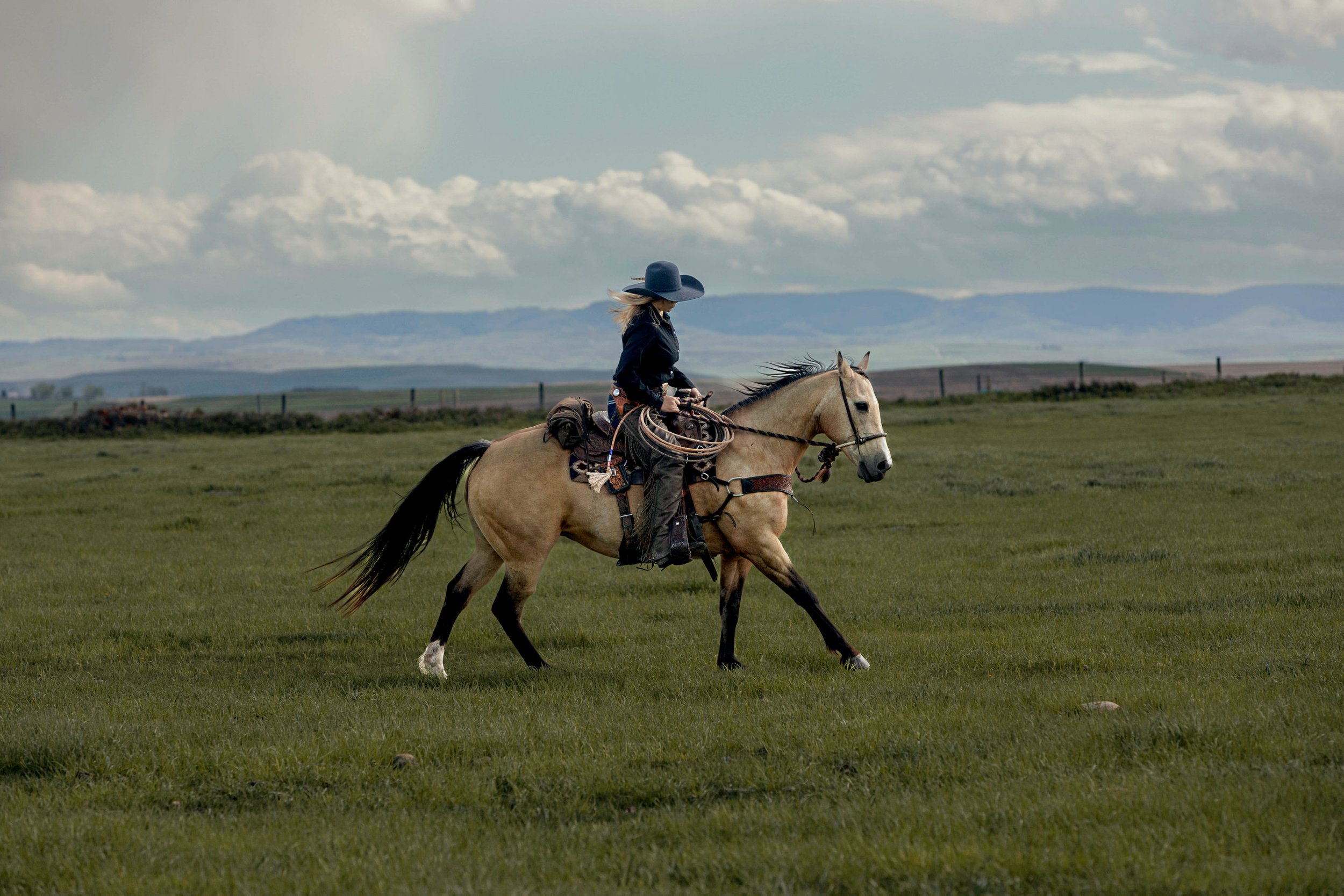 A woman riding a light-colored horse on a grassy plain with mountains in the distance and clouds in the sky.