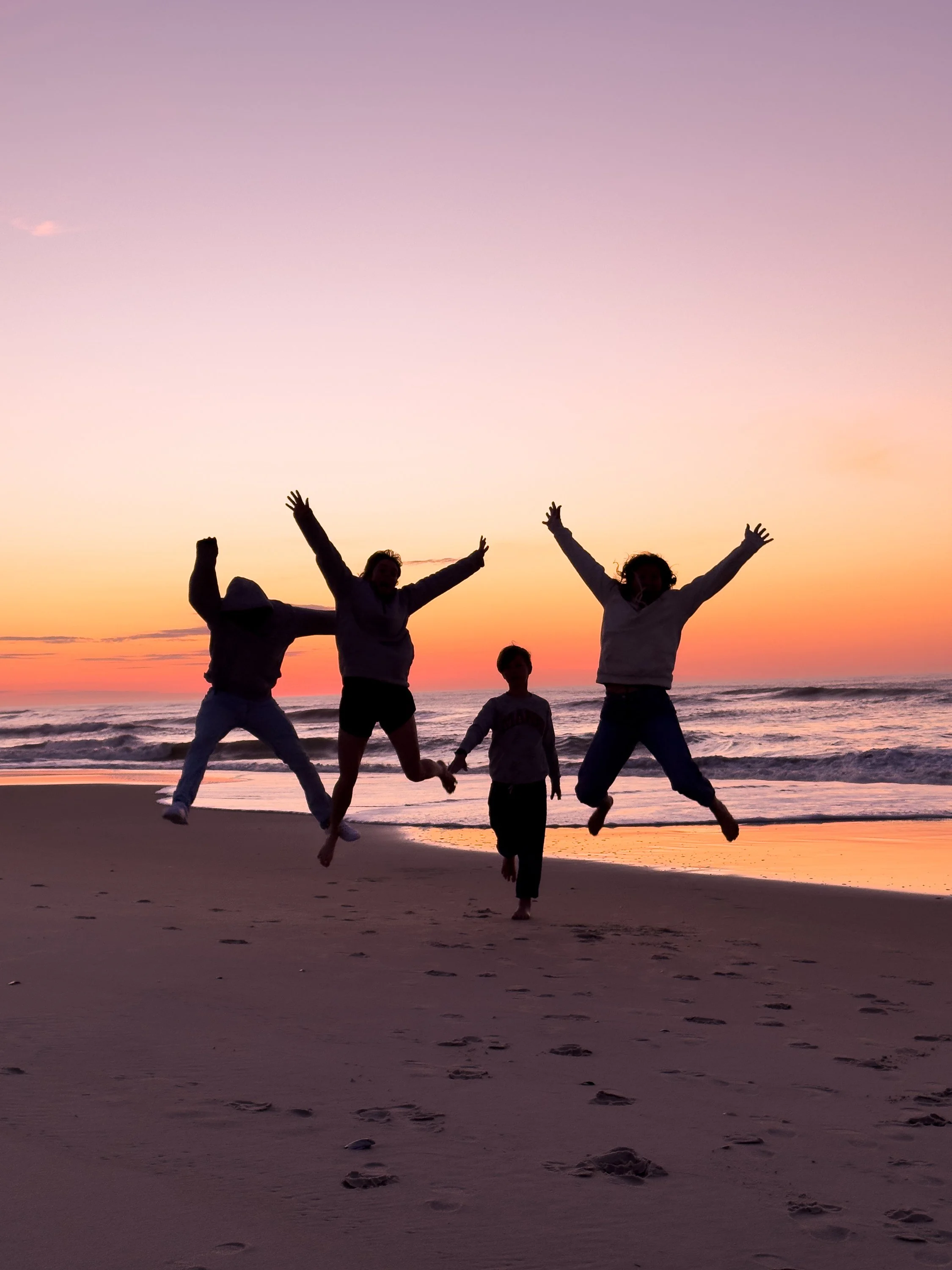 A group of people, including children and adults, are jumping and playing on a beach at sunset with colorful sky and ocean in the background.
