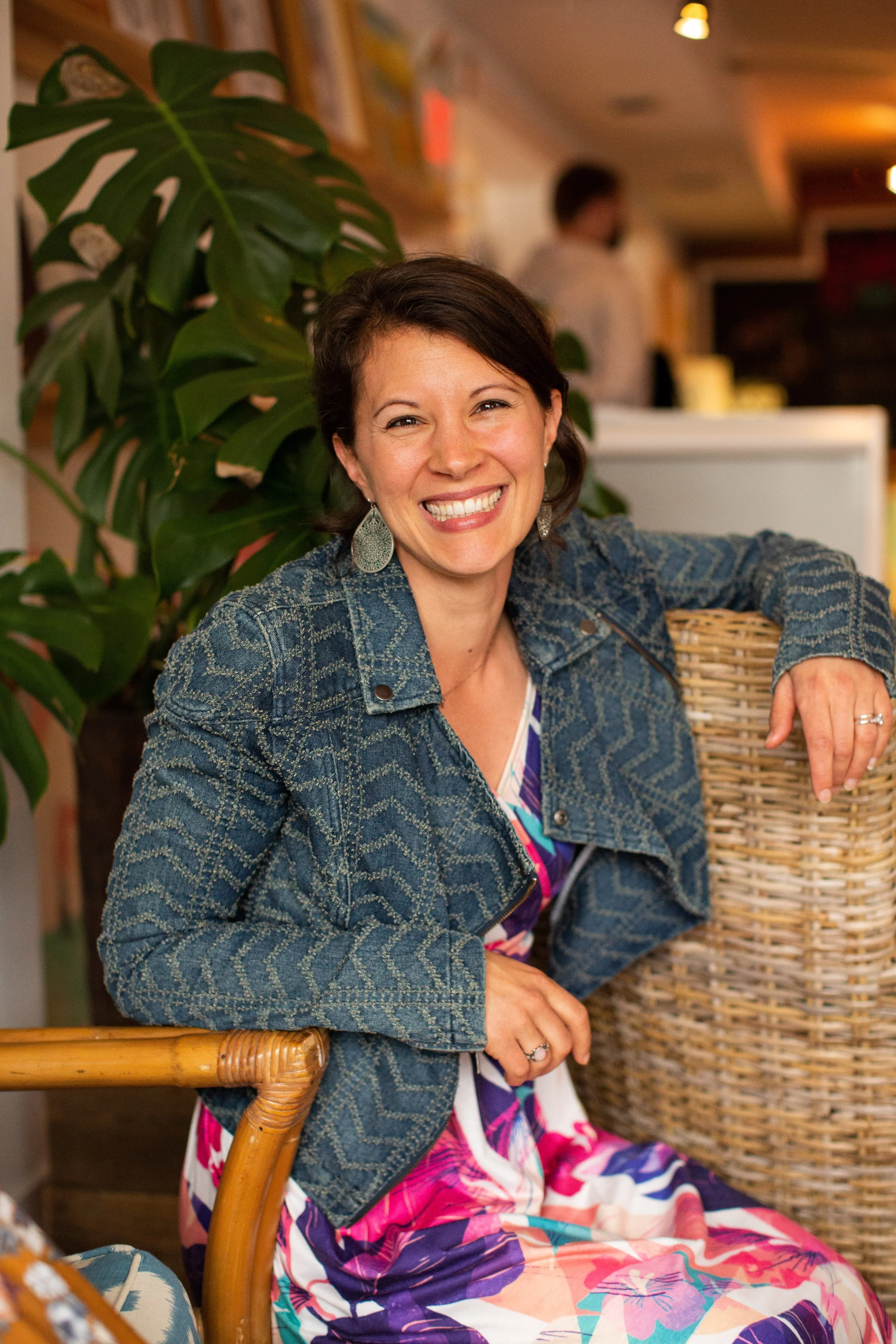 A woman sitting on a chair, smiling, wearing a patterned dress and a denim jacket with large earrings, with a large leafy plant behind her and a blurred figure in the background inside a warmly lit room.