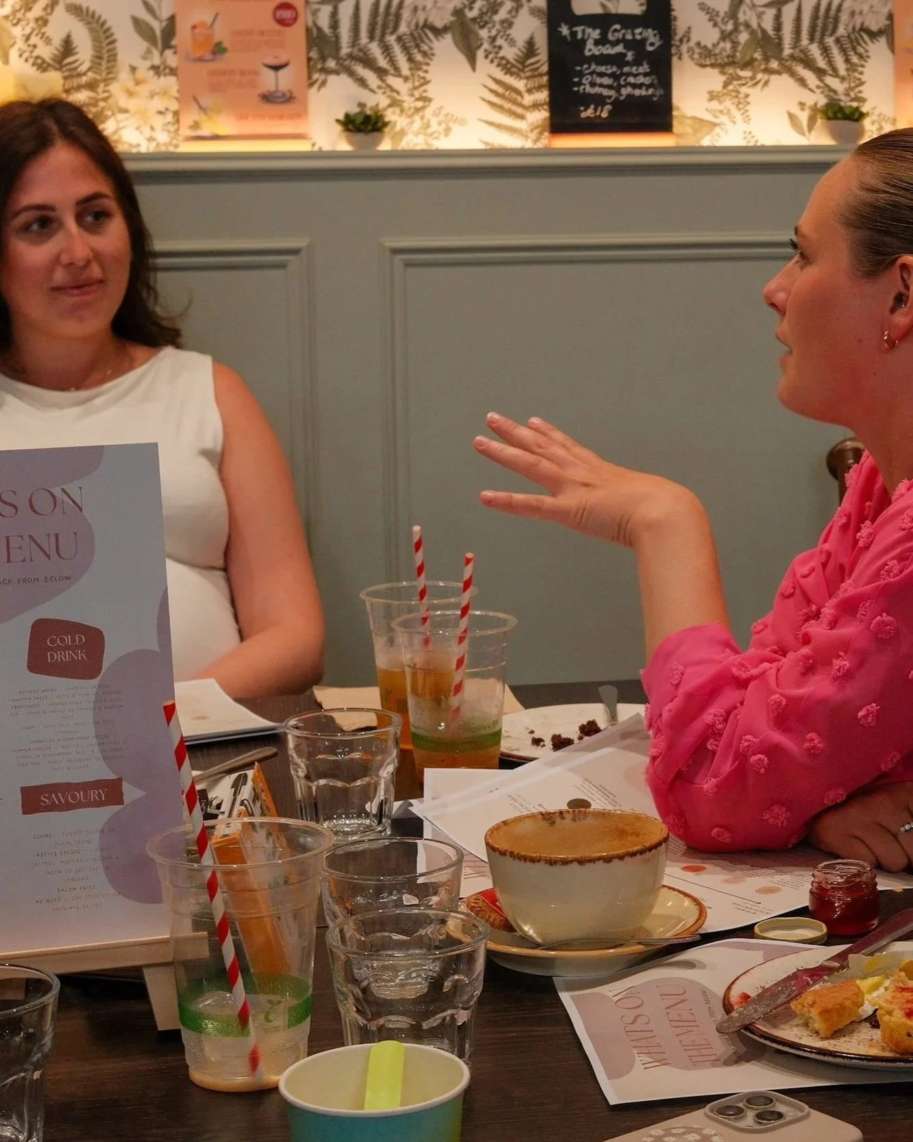 Two women are sitting at a dining table engaged in conversation. The table has drinks, plates with food, and menus. The background features patterned wallpaper and a blackboard menu.