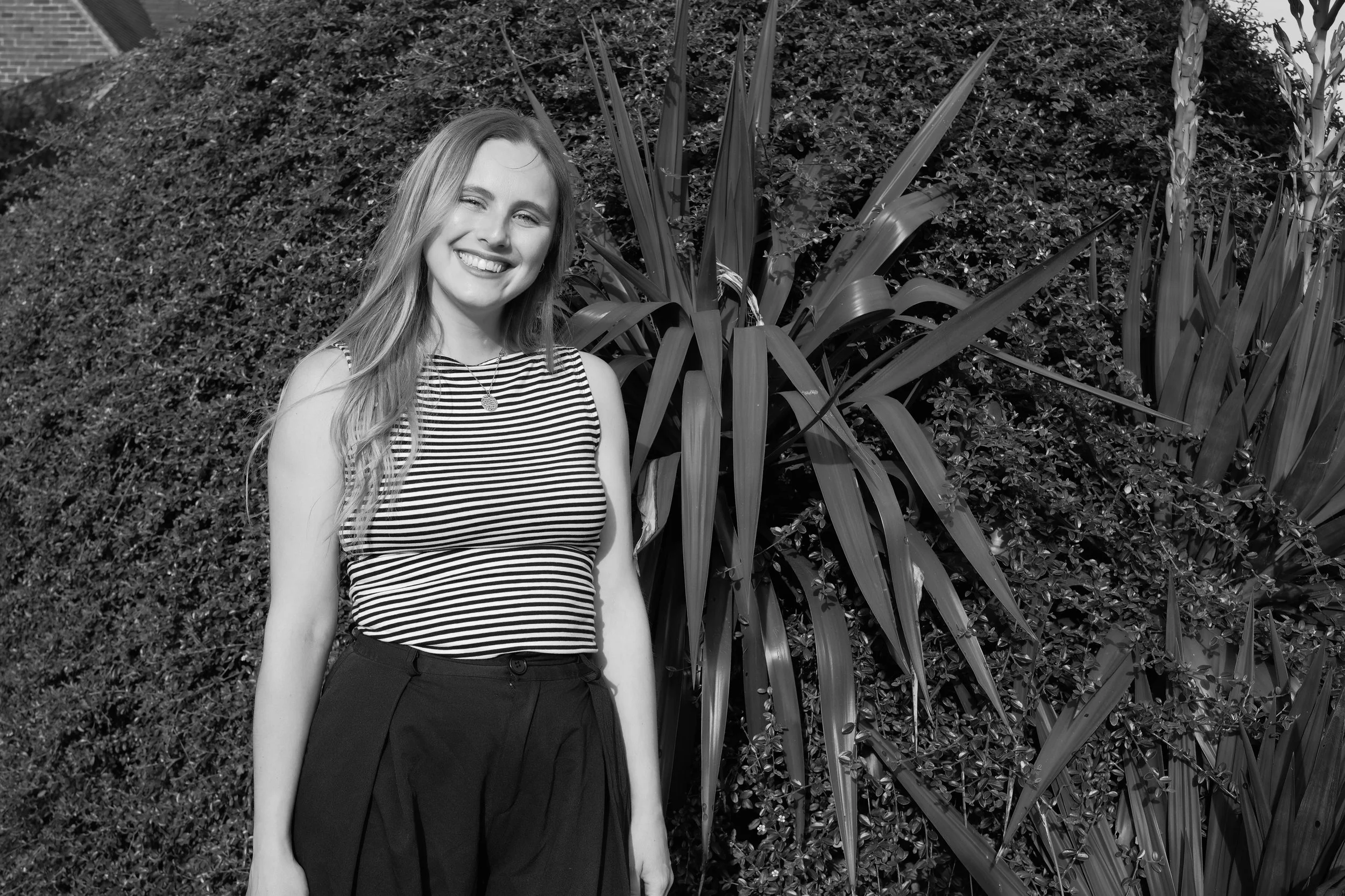 A young woman with long hair smiling and winking, standing outdoors in front of large plants and bushes, wearing a striped sleeveless top and dark pants, in black and white.