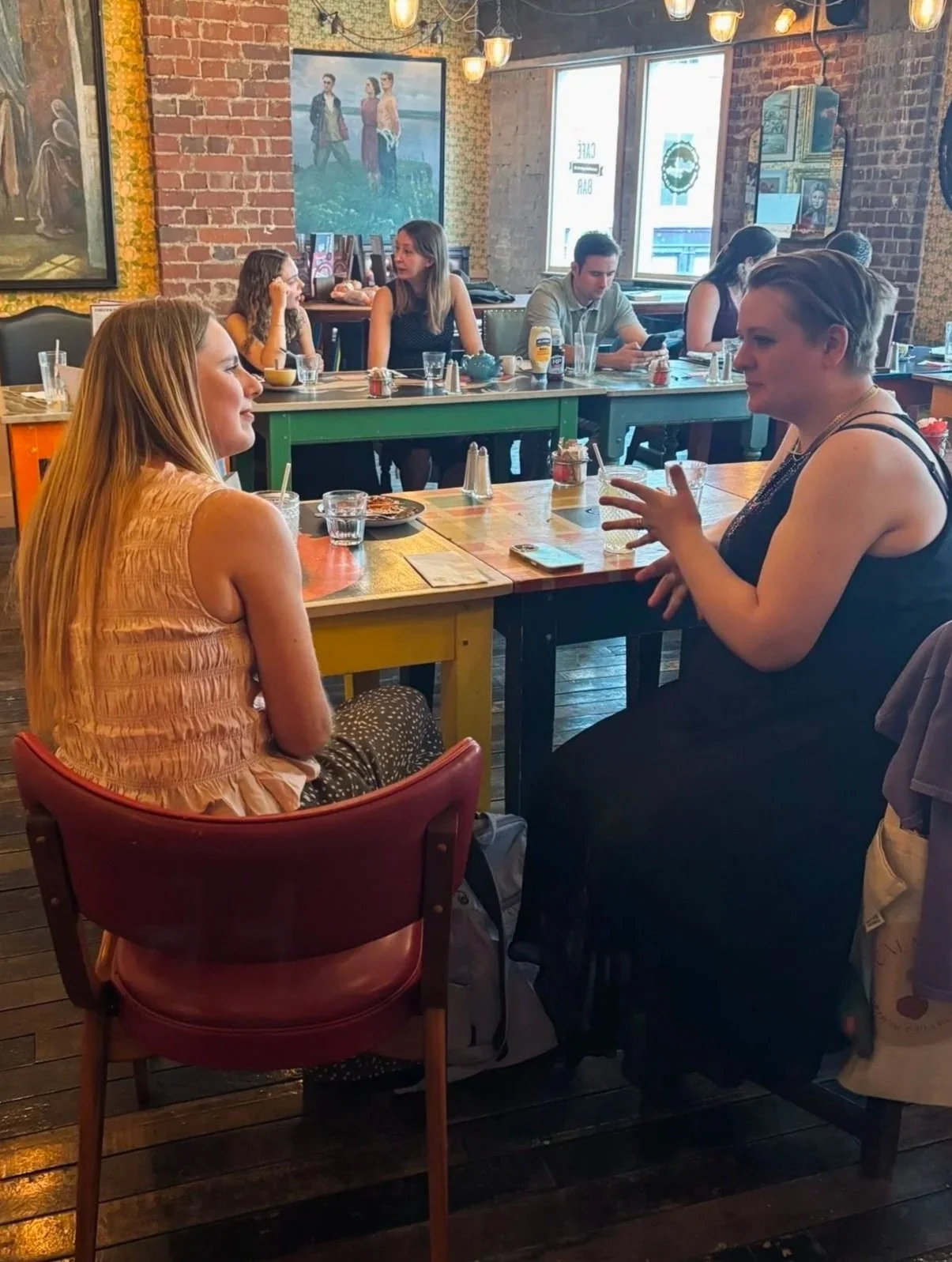 People having a conversation at a restaurant with exposed brick walls, paintings, and large windows, with a casual and lively atmosphere.
