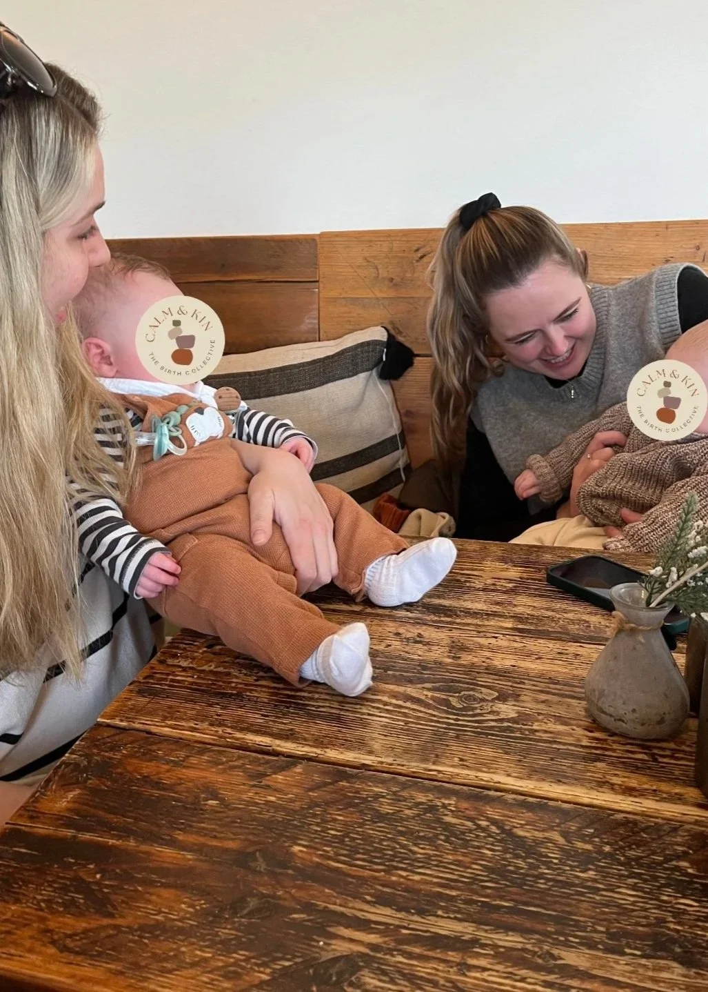 Two young women, one with long blonde hair and the other with long brown hair in a ponytail, are sitting at a rustic wooden table with a baby. The woman with blonde hair is holding the baby, who is dressed in orange and striped clothing, while the woman with brown hair is breastfeeding the baby. The scene appears warm and joyful, with a flower vase on the table.