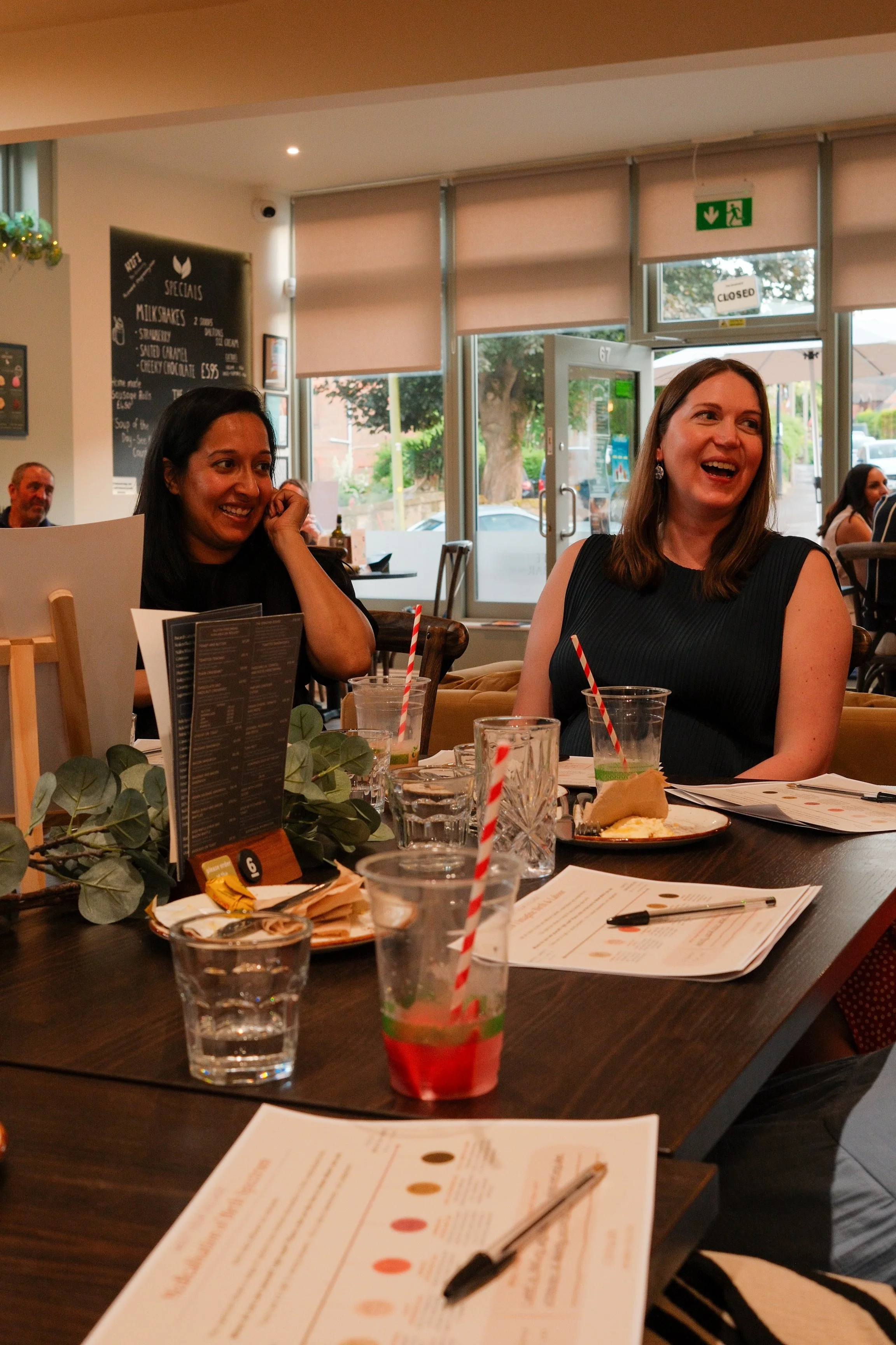 Two women sitting at a dining table, smiling and laughing. The table has drinks with straws, menus, and plates of food. The background shows a restaurant interior with large windows and other diners.