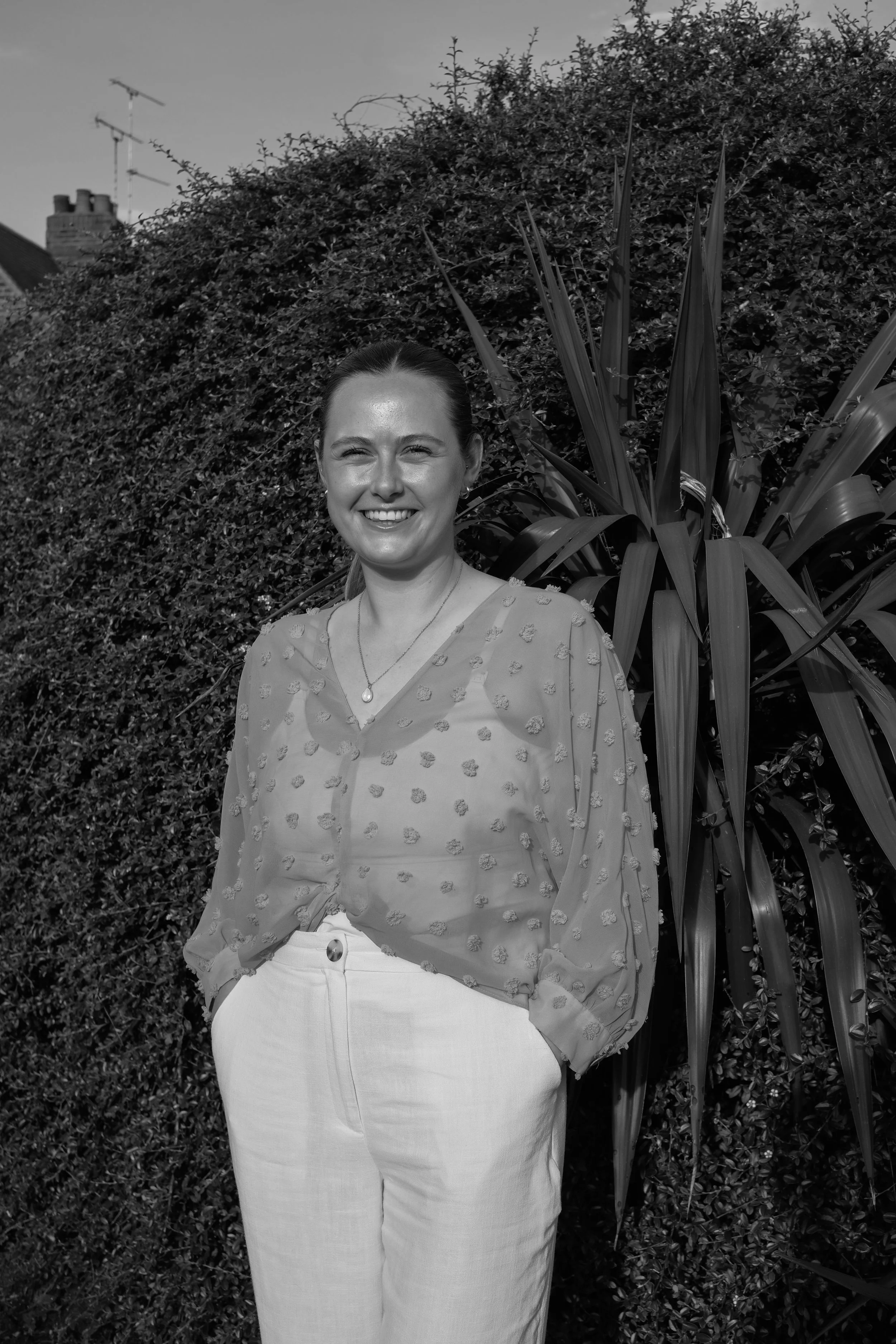 A woman smiling outdoors, standing in front of a large bush and a tall plant, with rooftops and chimneys in the background, captured in black and white.