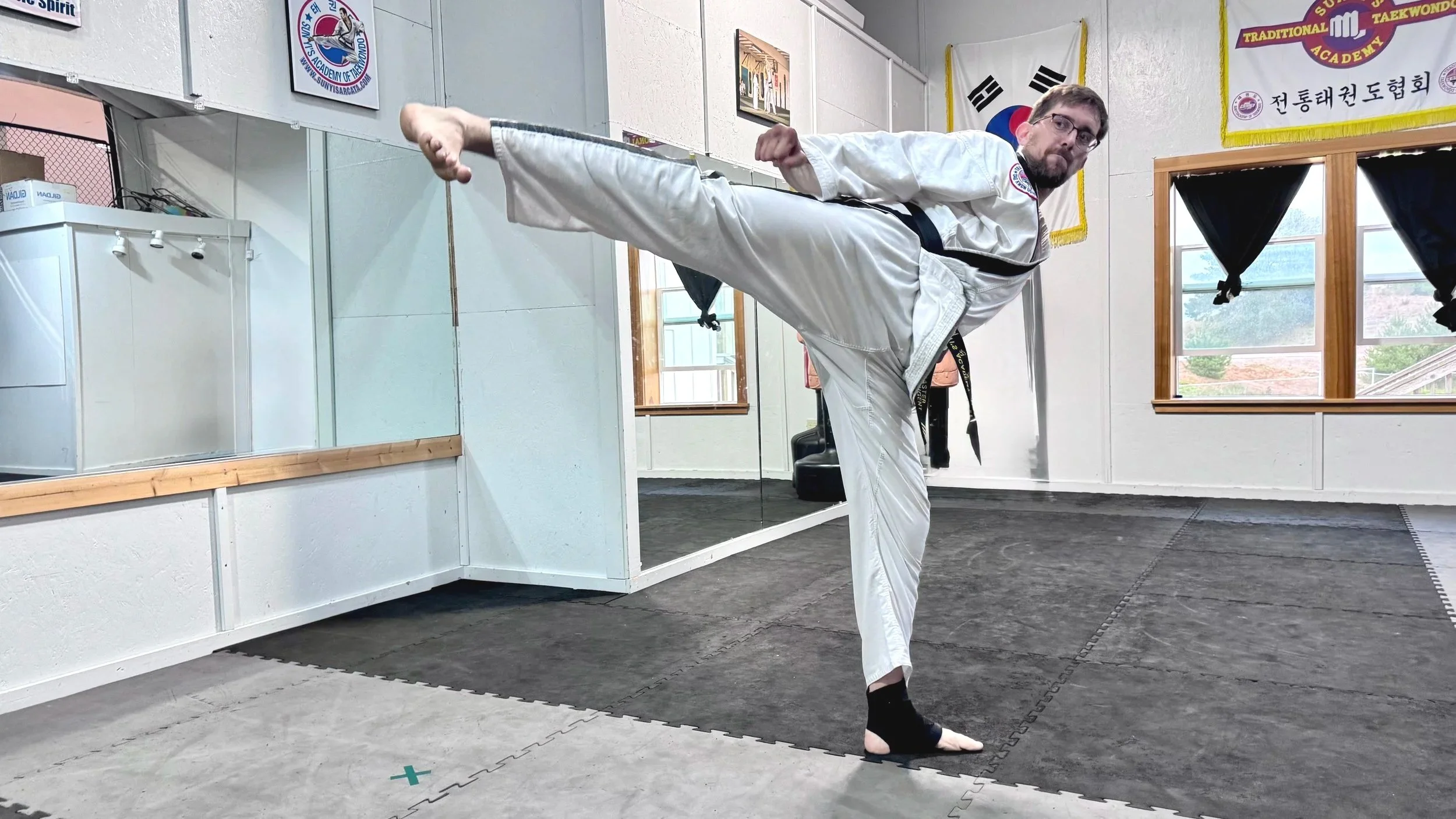 A man in a white martial arts uniform demonstrating a high kick in a martial arts studio, with flags and martial arts banners on the walls, and a mirror reflecting part of the room.