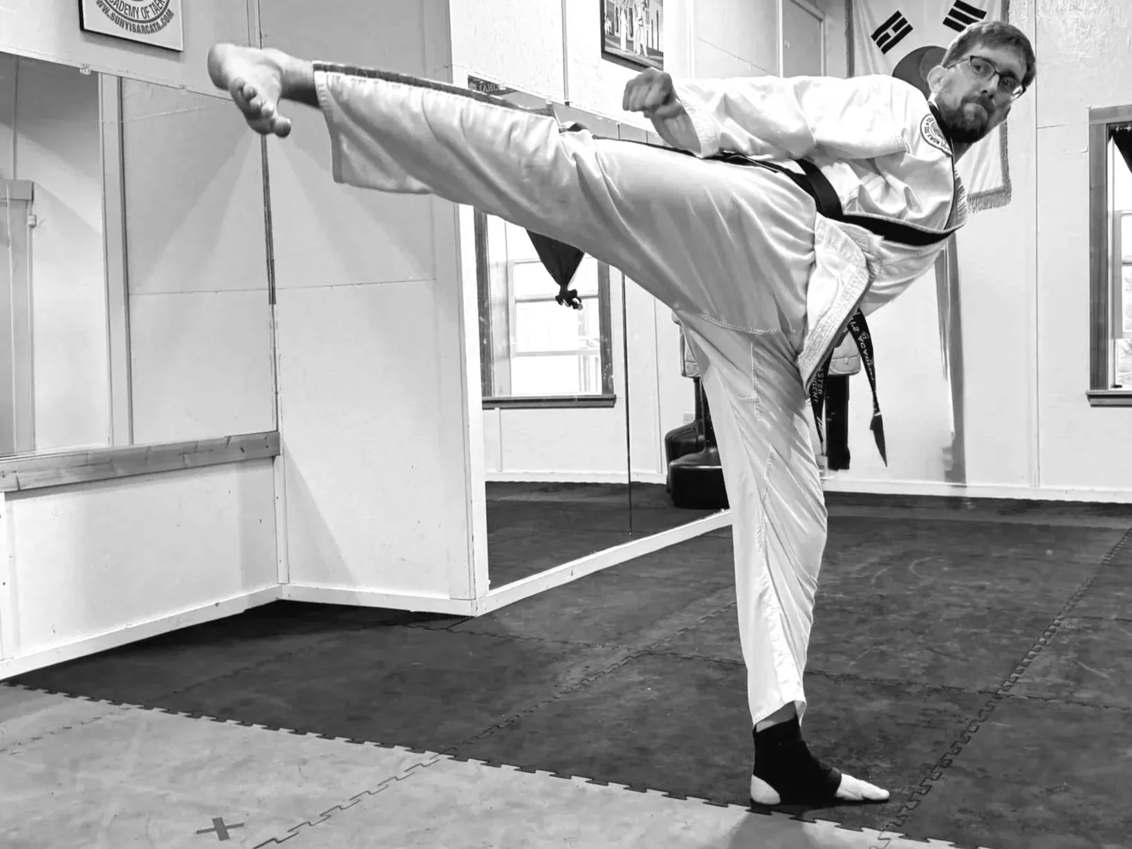 A man practicing martial arts in a gym, performing a high side kick while wearing a martial arts uniform, black belt, and glasses.