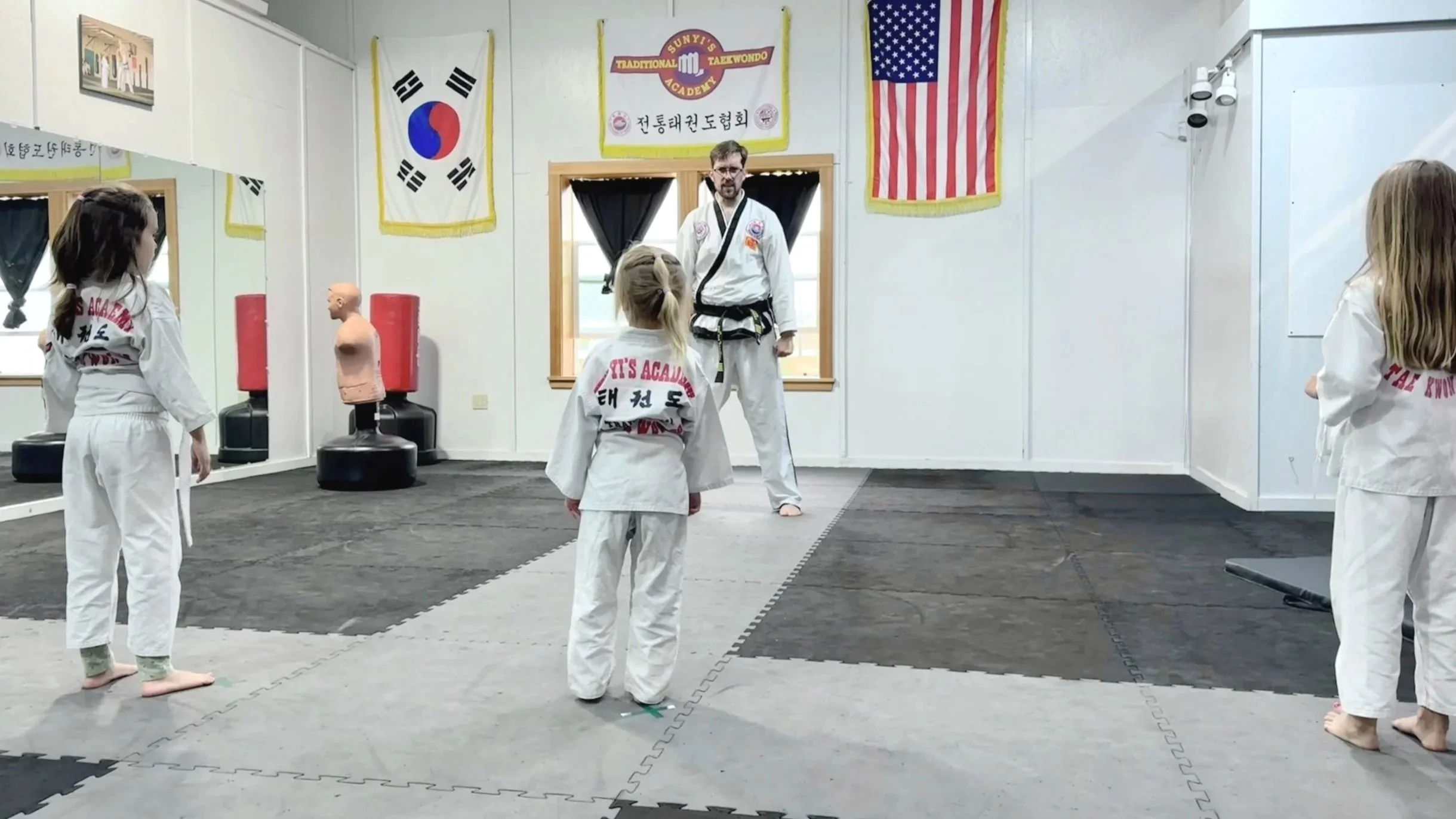 Martial arts class in a dojo with students in white uniforms and instructor, Korean and American flags hanging on the wall, training equipment visible.