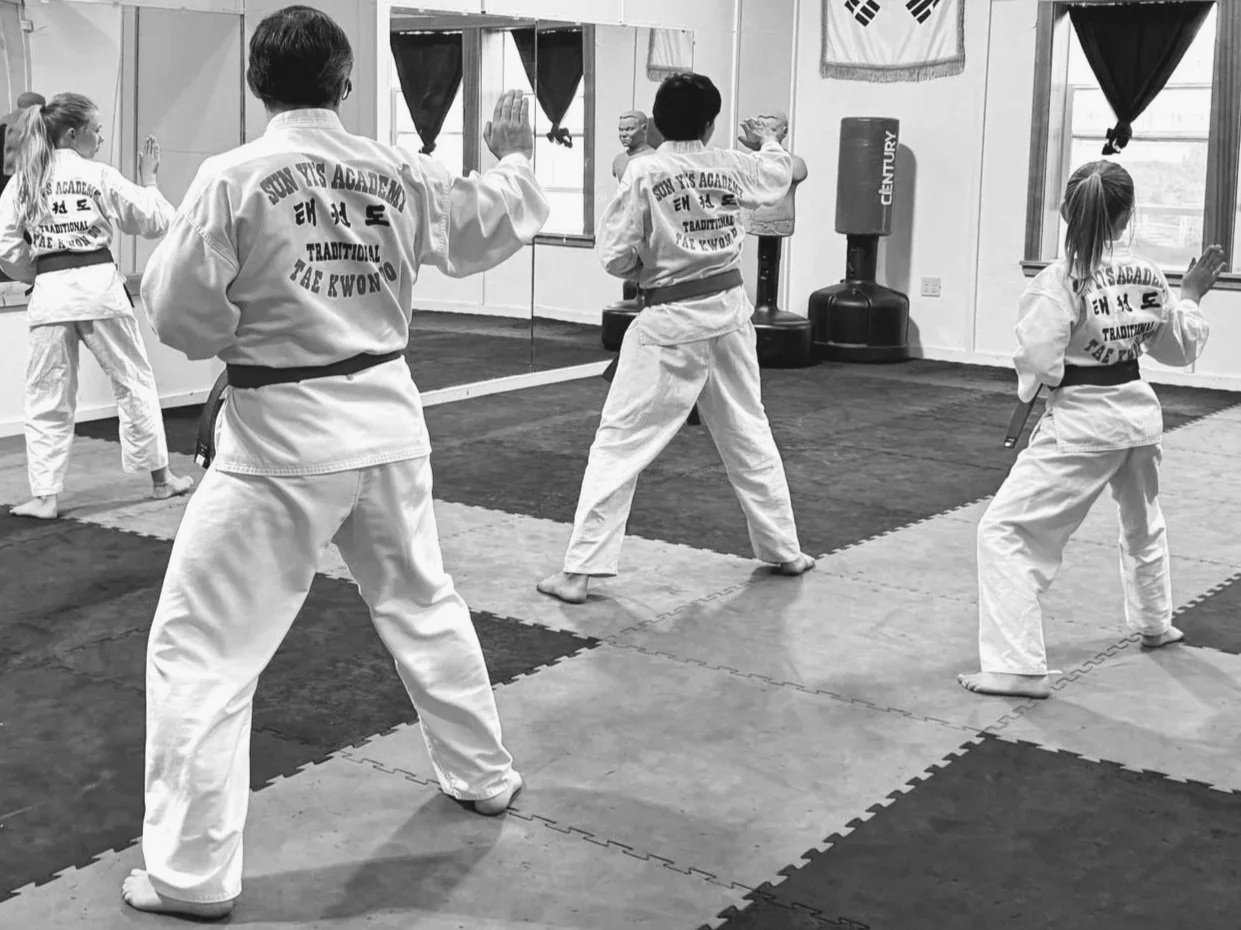 Children practicing martial arts in a dojo, wearing uniforms with 'Sun Yi's Academy' on the back, and an instructor observing in front of a mirror.