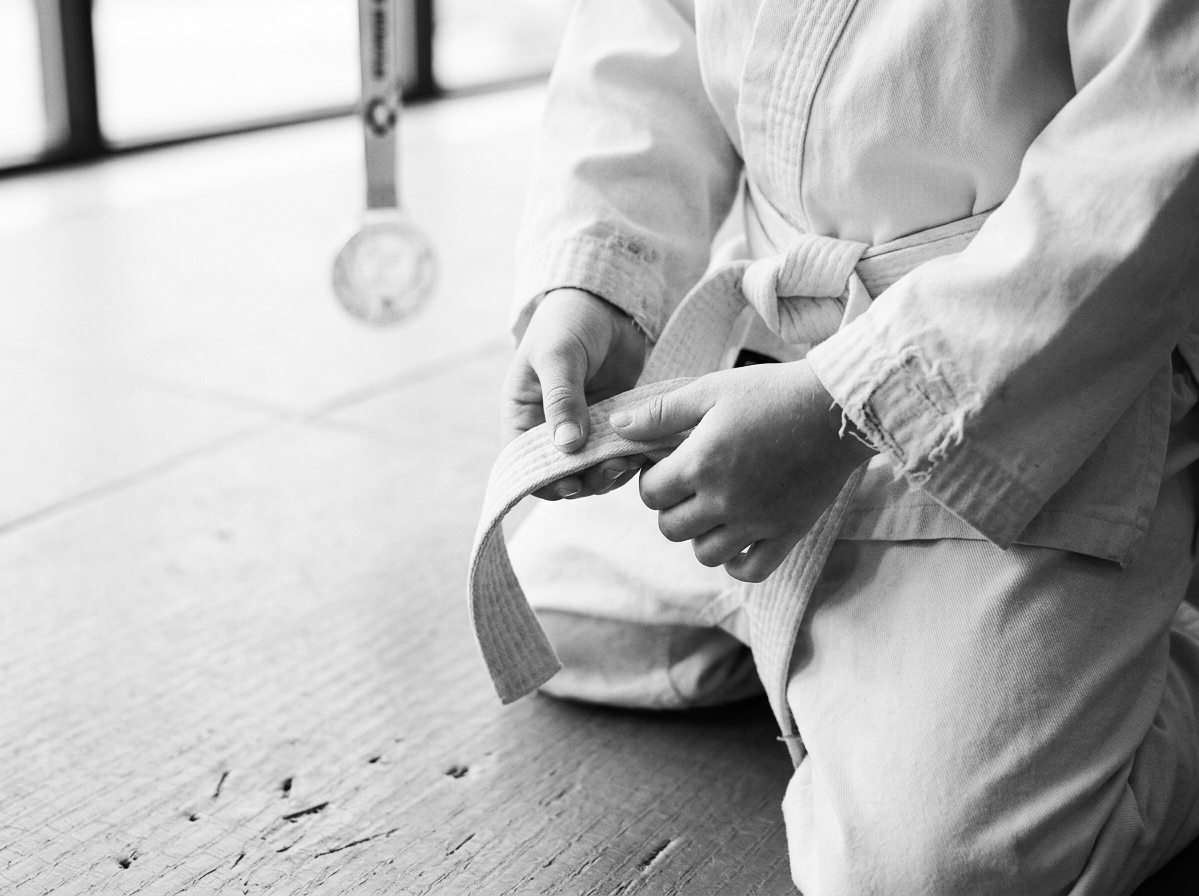 A person wearing a martial arts gi tying their belt, kneeling on a tatami mat in a dojo.