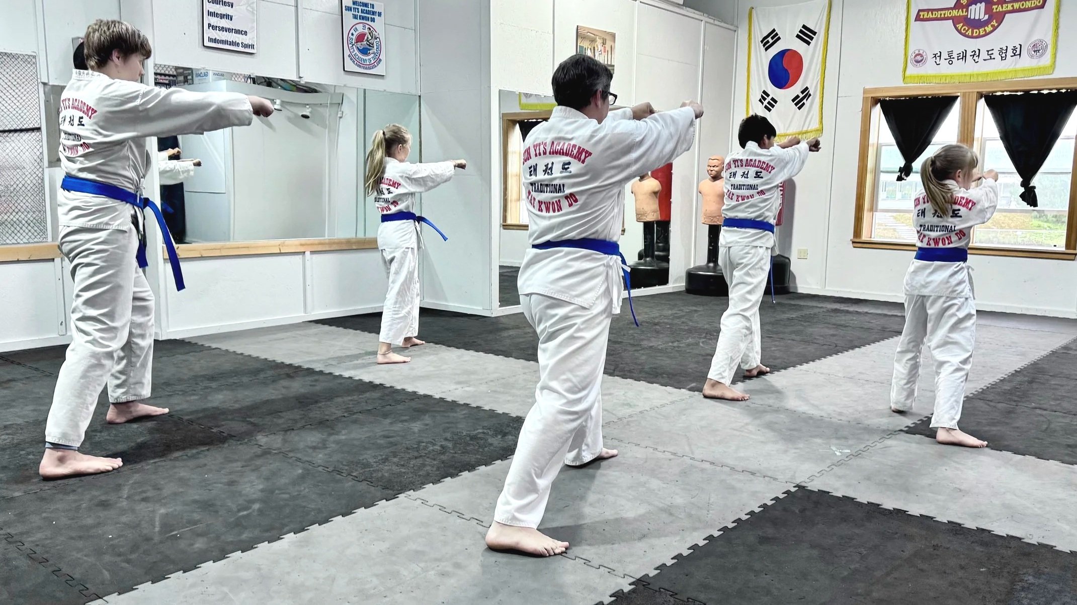 Martial arts class with children practicing techniques in a dojo, wearing white uniforms and blue belts, with South Korean flags and martial arts banners hanging on the walls.