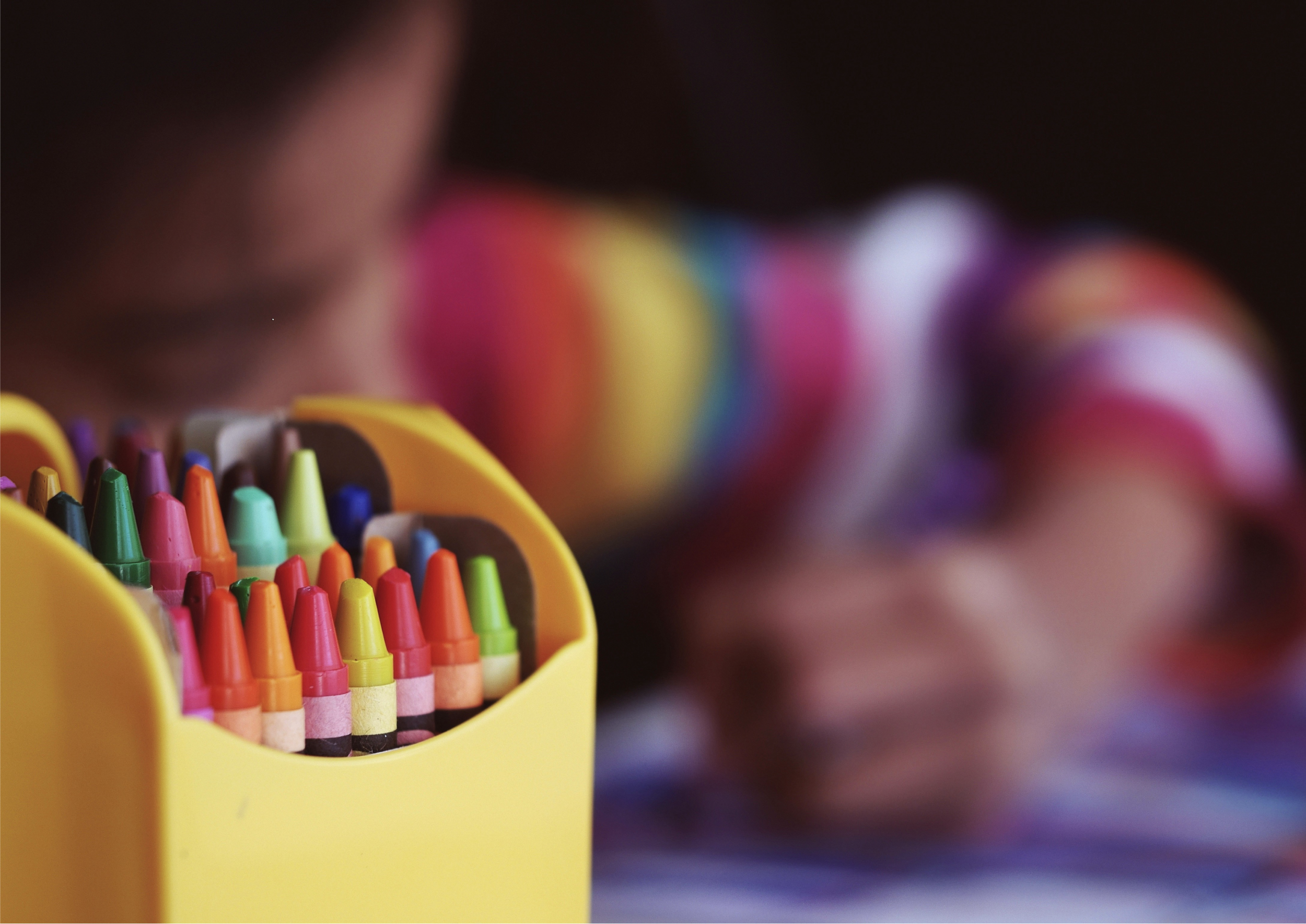 A yellow case containing a variety of colorful crayons, with other art supplies blurred in the background.