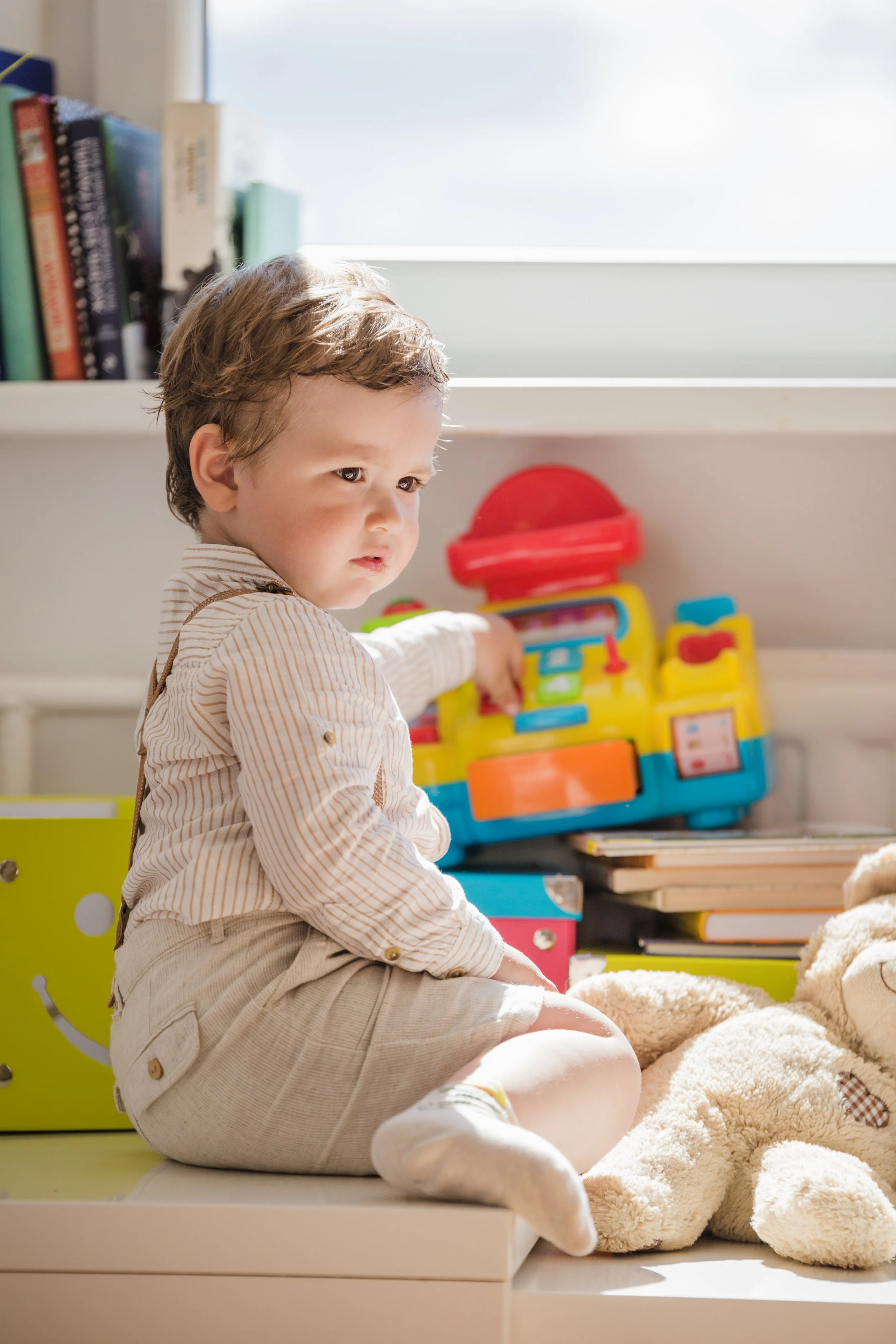 A young boy with light brown hair sits on a window seat, reaching toward colorful toys and a pile of beige stuffed animals. Bright sunlight streams through the window, illuminating a bookshelf and storage bins in the background.