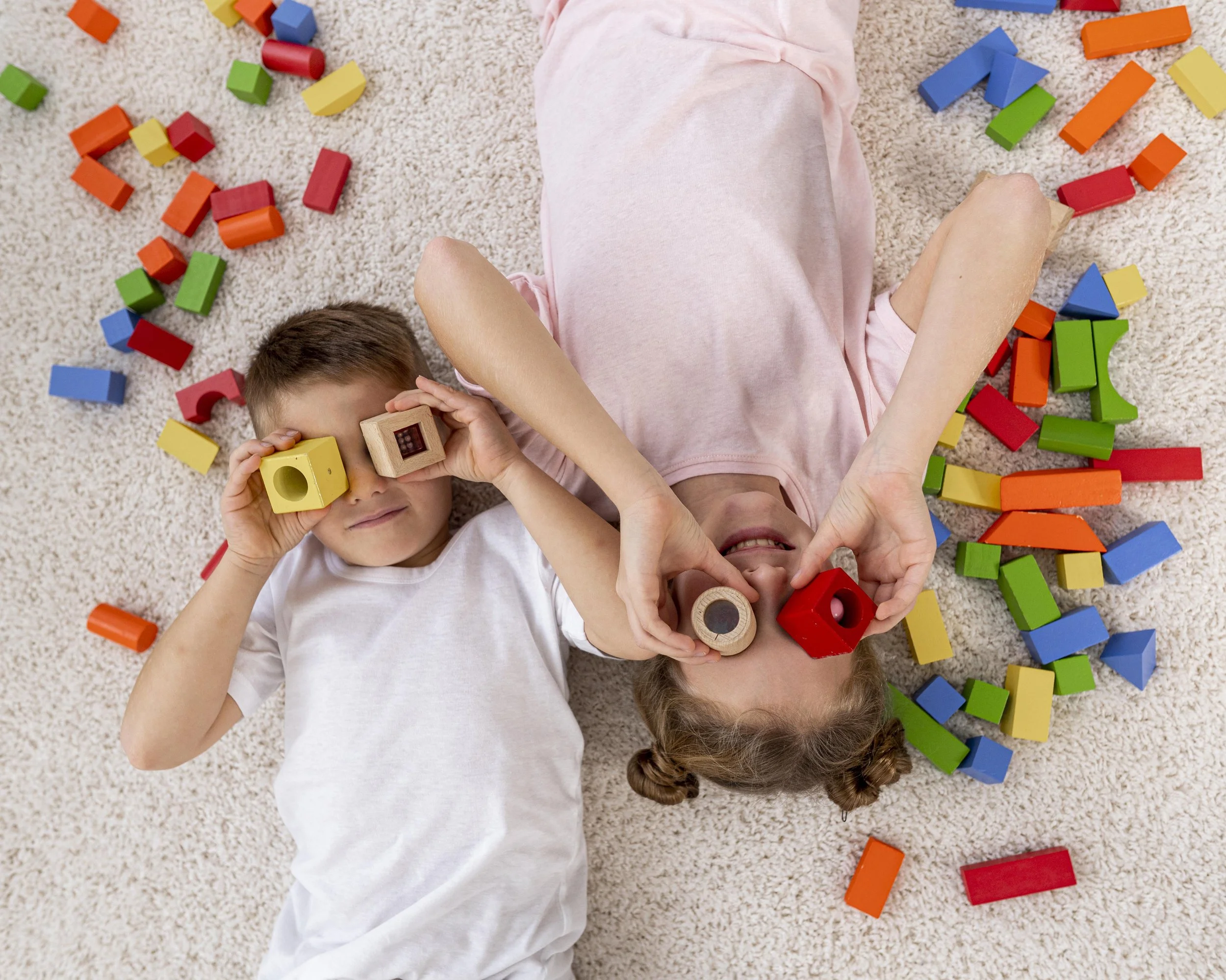 Three children lying on a beige carpet surrounded by colorful toy building blocks. One child is holding a wooden block up to their eye, another is making a heart shape with their hands, and the third is partially obscured.