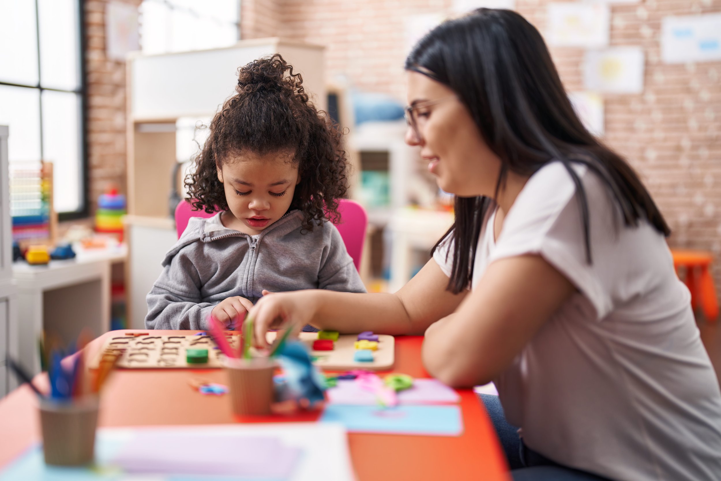 A young girl and an adult woman sitting at a table engaged in a craft activity, surrounded by colorful materials in a room with brick walls and large windows.