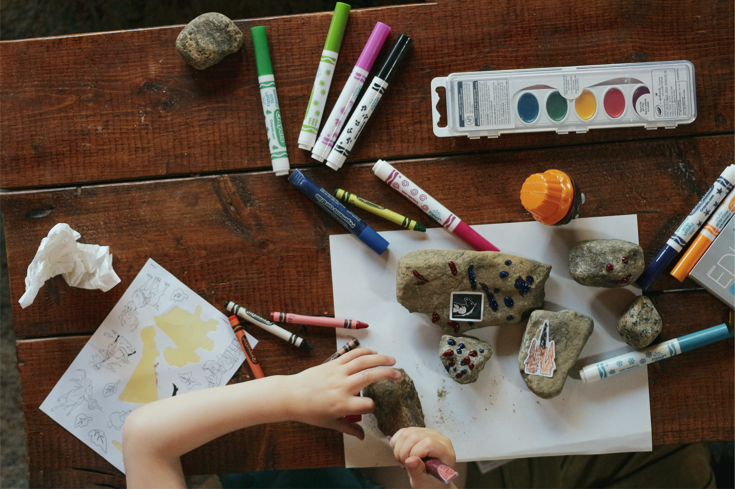 Child's hand organizing painted rocks with small stickers on a wooden table, surrounded by markers, drawings, and a paint set.