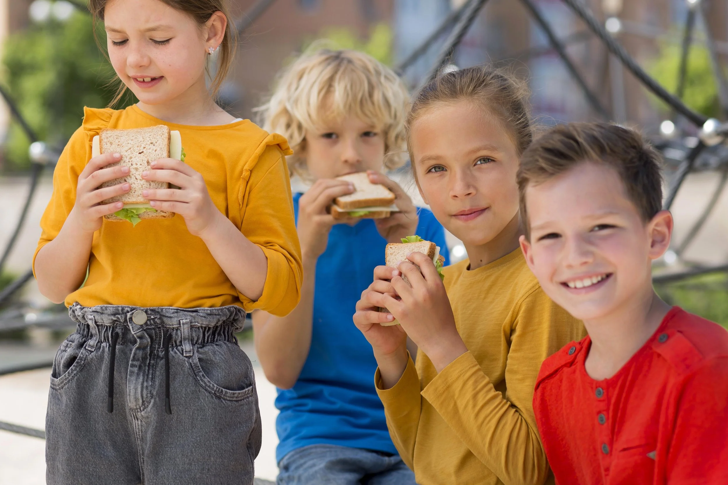 Four children sitting outdoors, eating sandwiches, with a playground structure in the background.