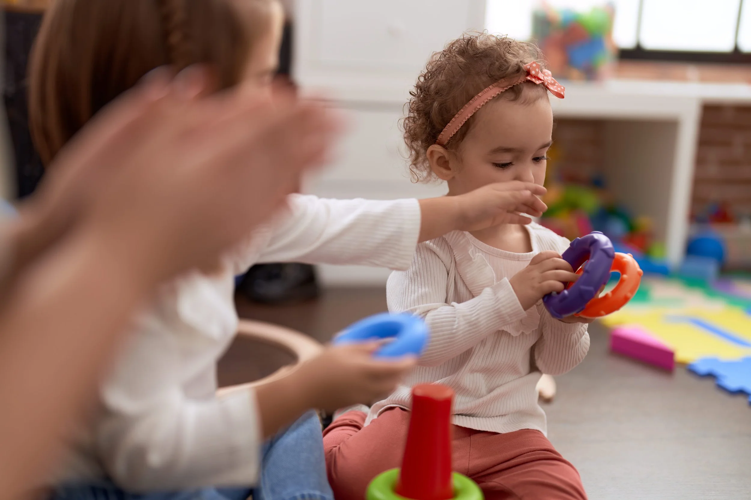 Young girl with curly hair and a pink polka-dot headband playing with colorful toys in a playroom.