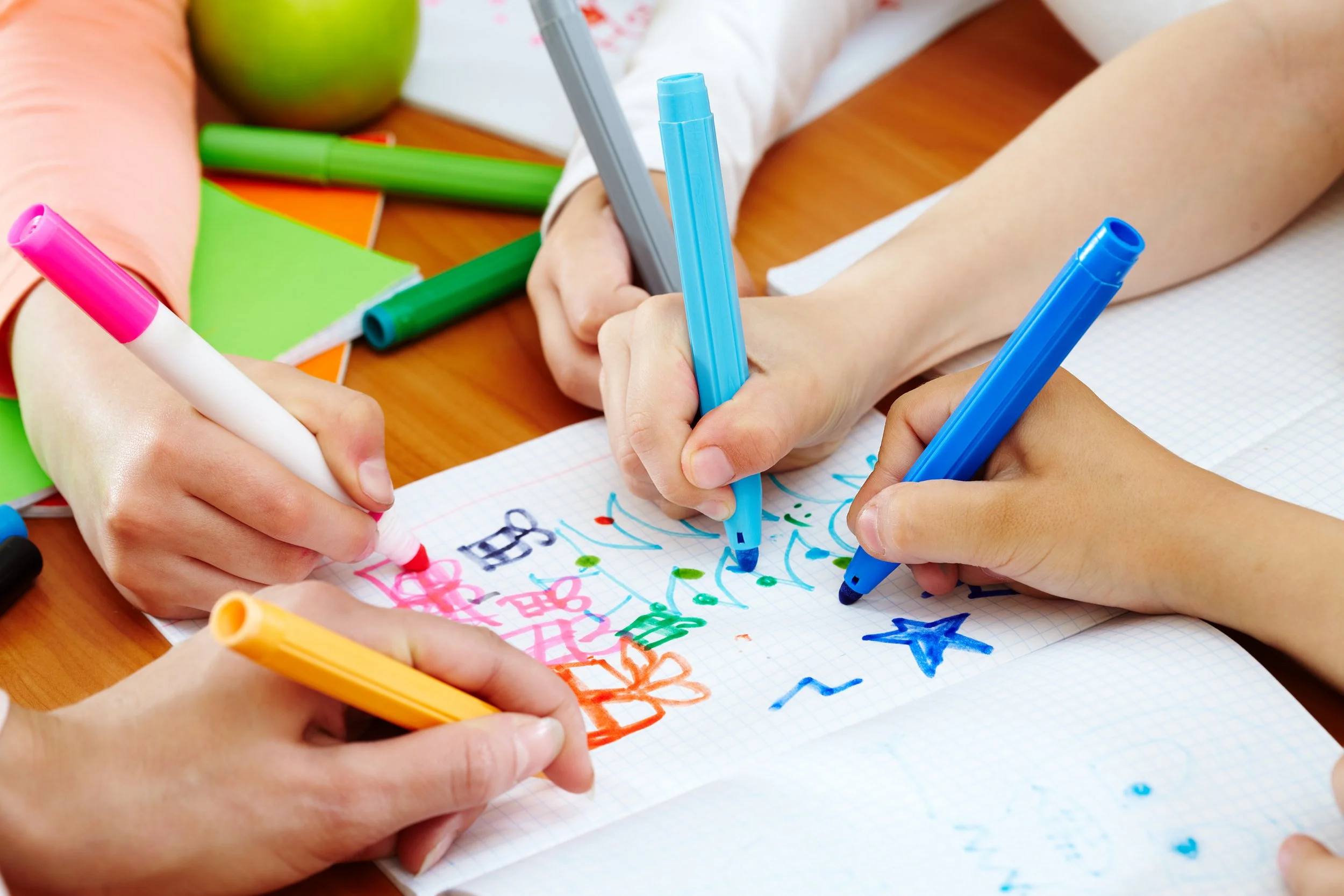 Close-up of multiple hands holding colorful markers, writing and drawing on a sheet of graph paper. Various colored markers and papers scattered around on a wooden surface.