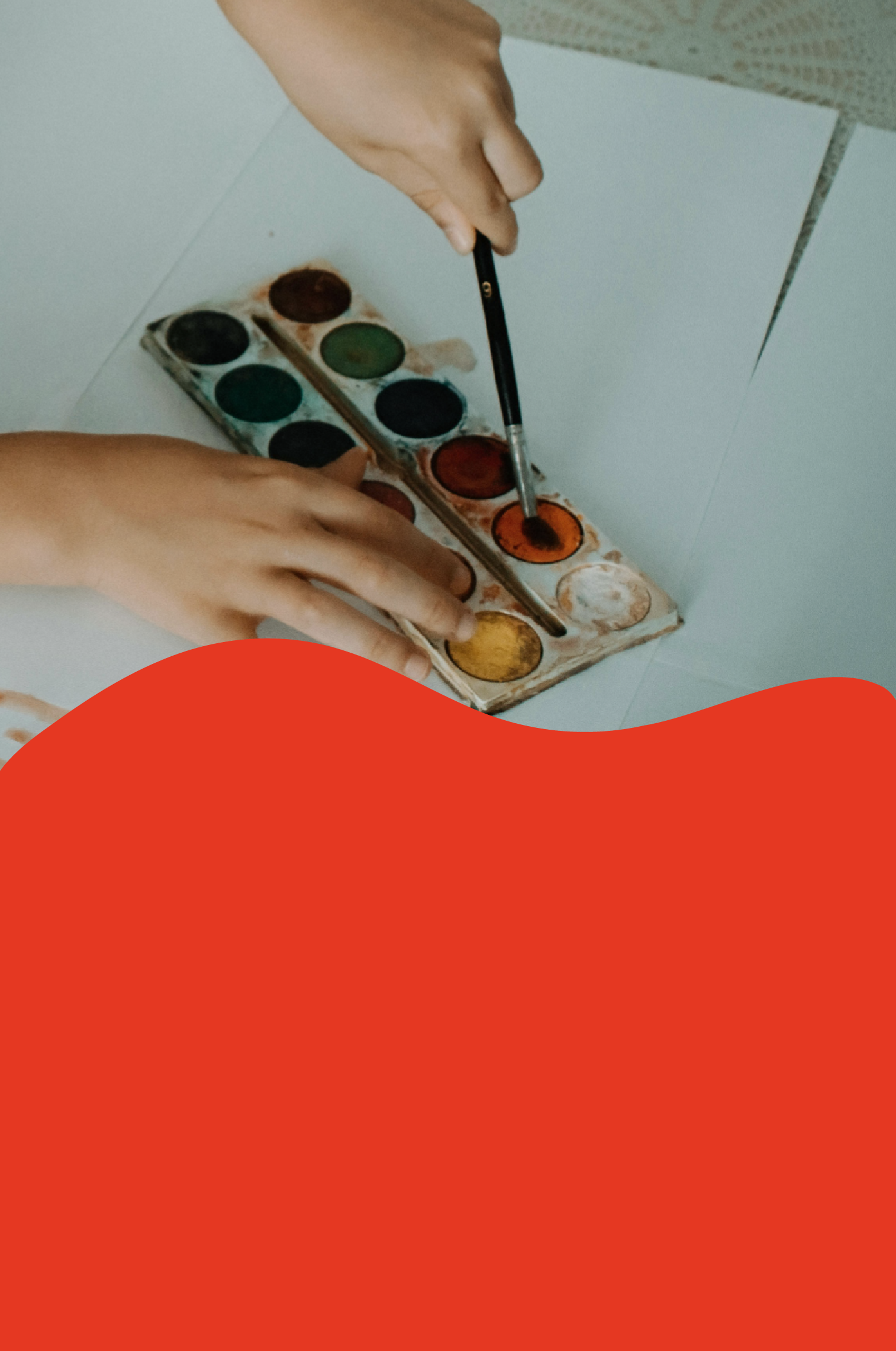 Hands holding a watercolor paint palette with various colors, and a paintbrush dipping into orange paint.