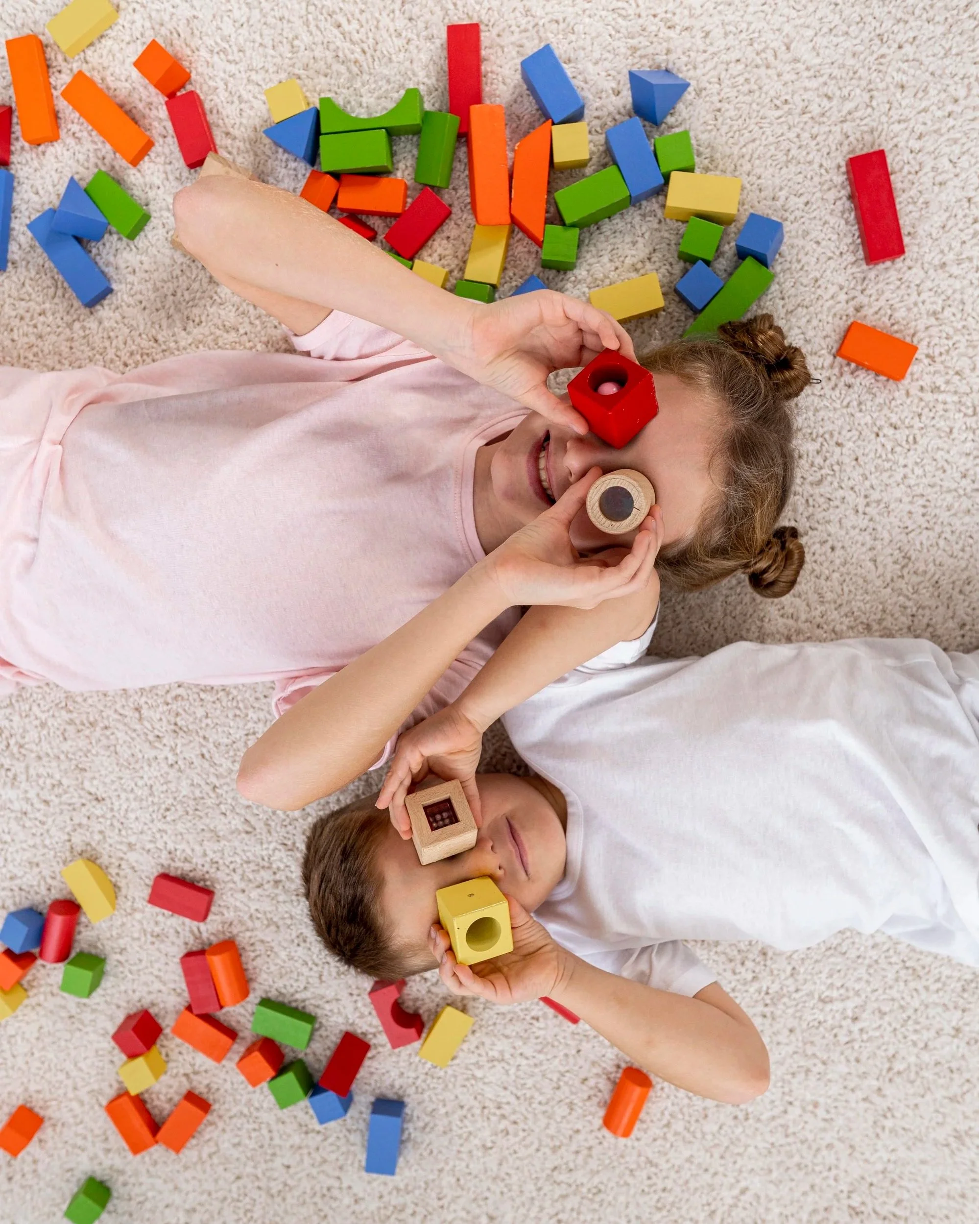 Children lying on carpet surrounded by colorful wooden blocks, some children hold blocks up to their eyes.