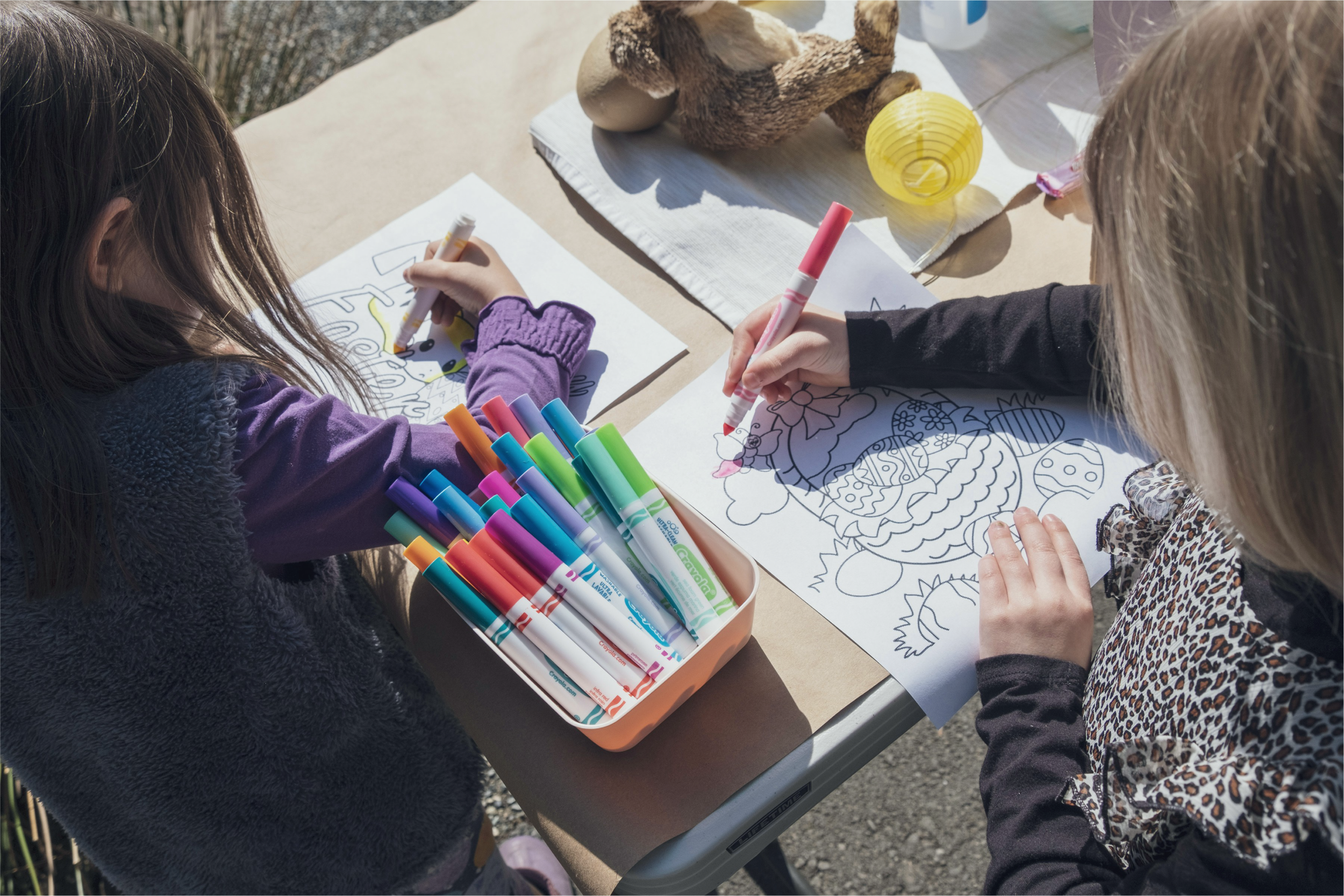 Three children drawing and coloring on paper at a table outdoors with art supplies and decorative objects.