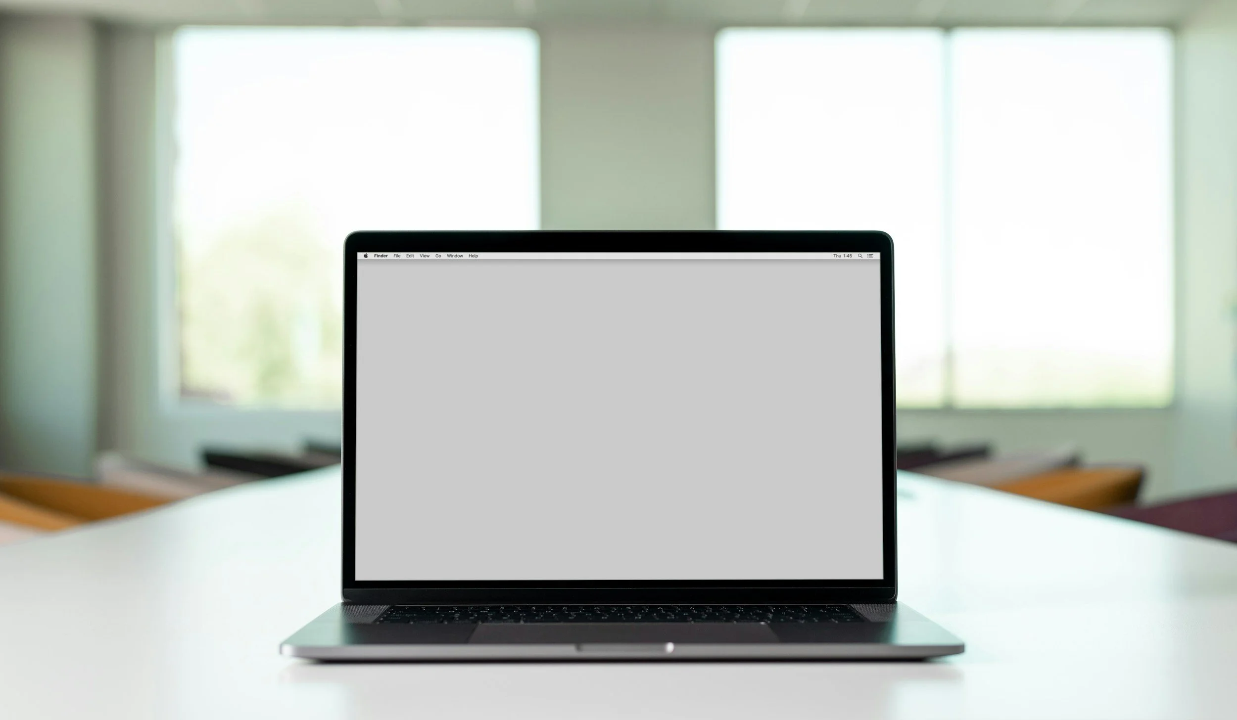 Laptop with blank gray screen on a white table in front of large windows with closed blinds.