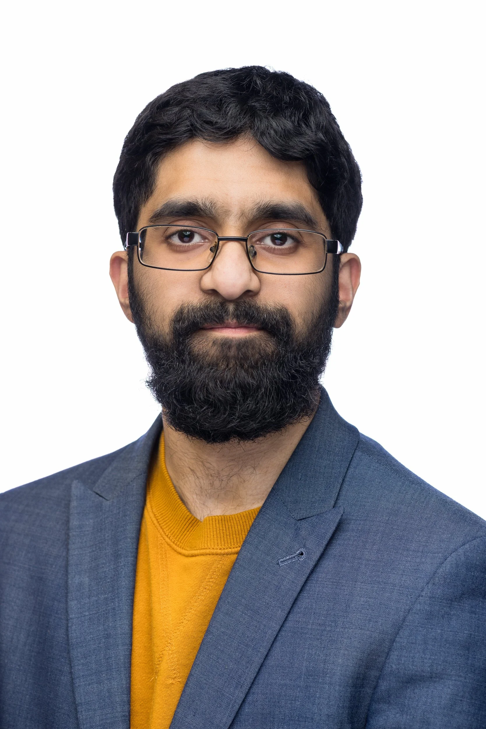 A portrait of a man with black hair, glasses, and a beard, wearing a gray blazer over a mustard yellow sweater.