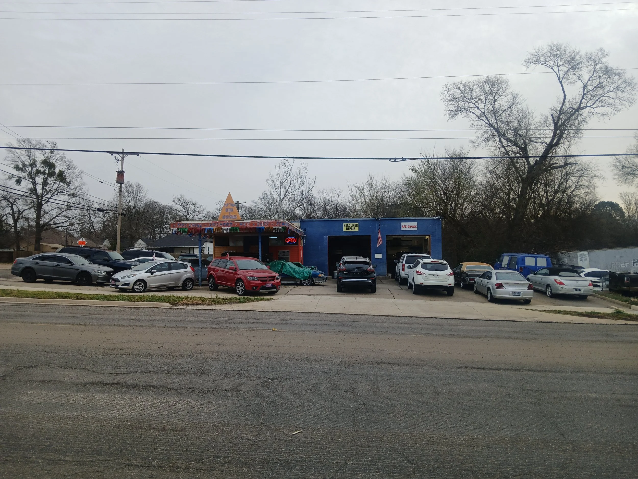 A parking lot with various cars in front of a blue building that has signs indicating auto repair services. There are trees without leaves in the background and power lines overhead on an overcast day.