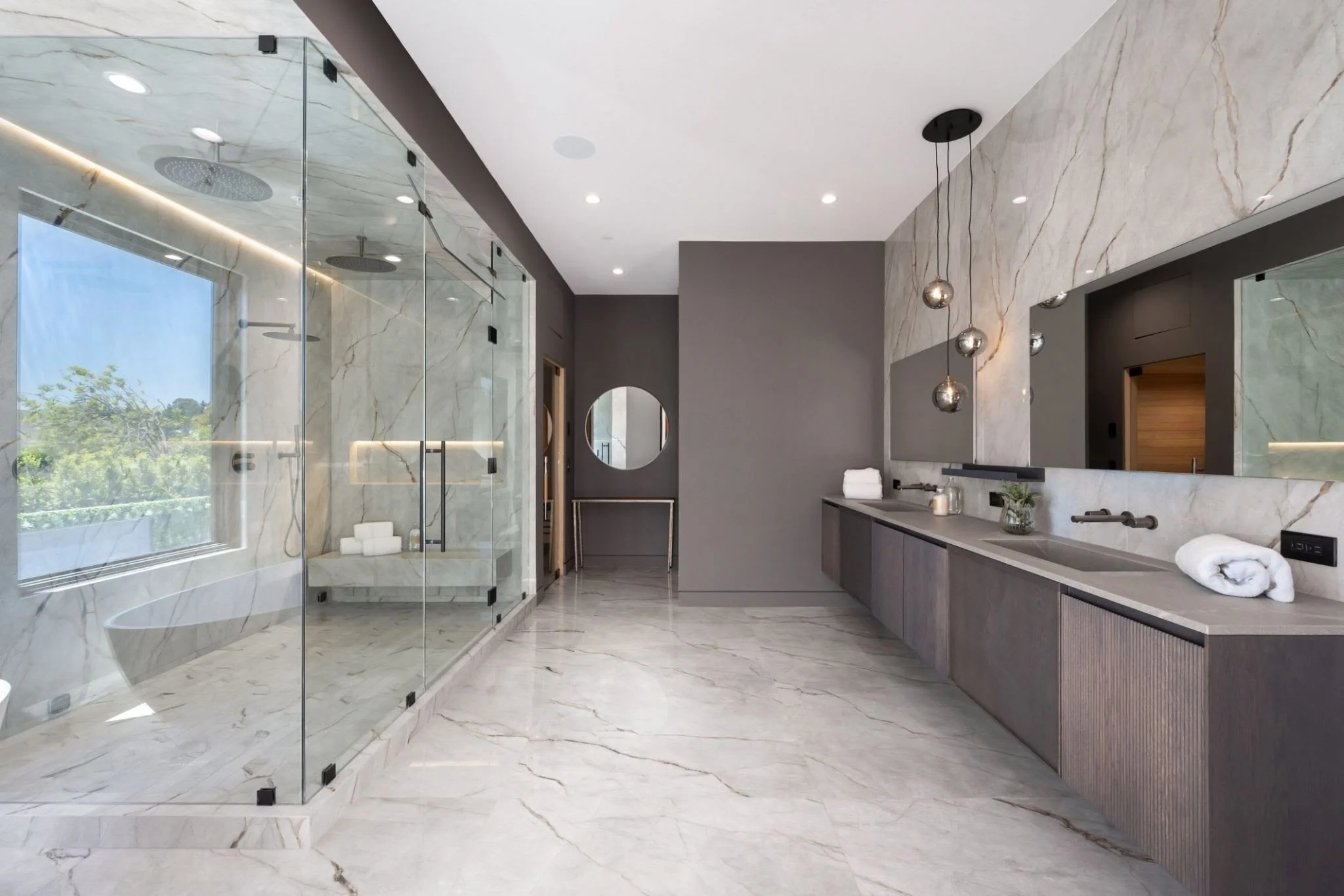 Modern bathroom with marble floors and walls, a glass-enclosed shower with rainfall showerheads, a vanity with a large mirror, and contemporary pendant lights.