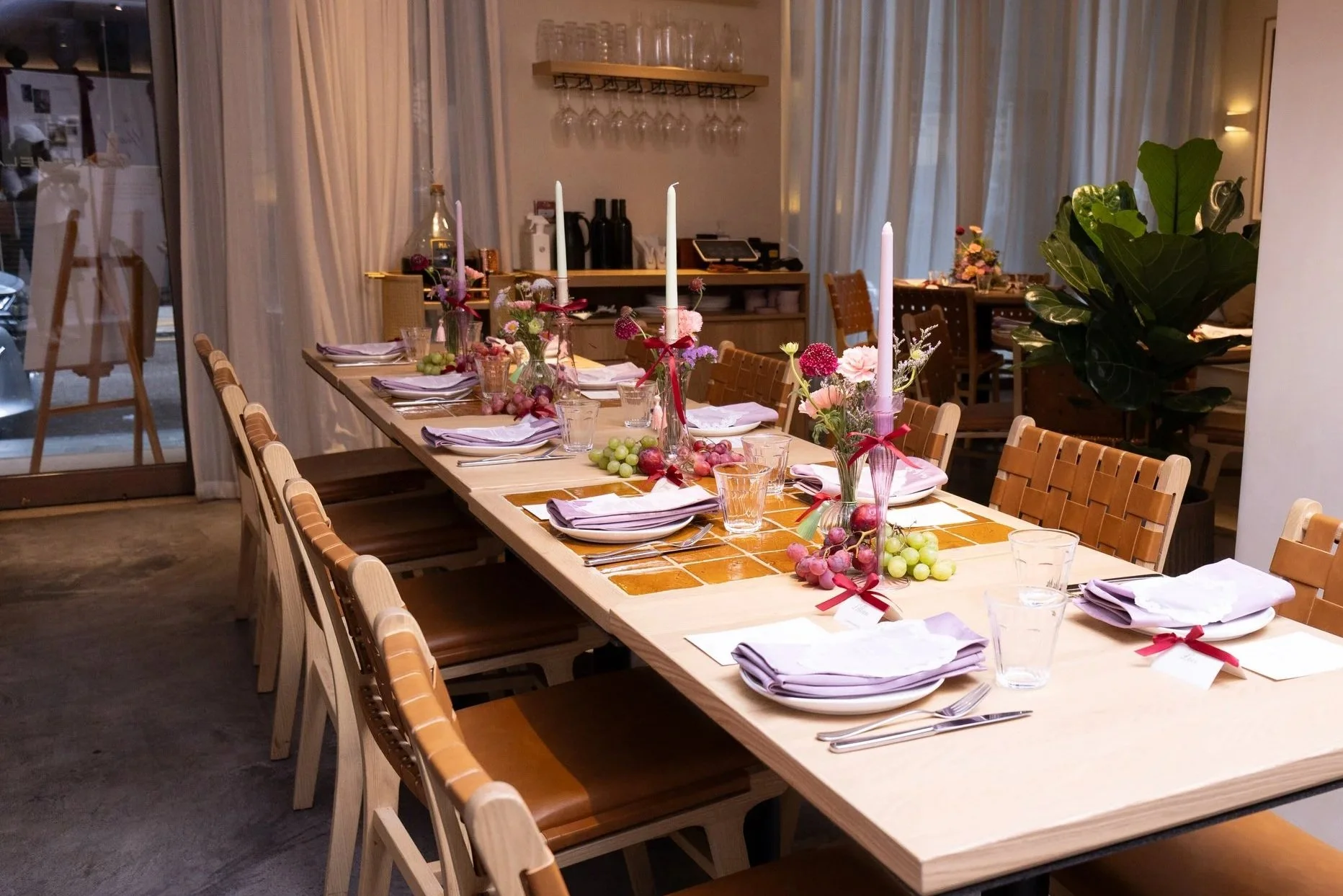 A long wooden dining table set for a meal with pink flower centerpieces, candles, glasses, plates, napkins, and cutlery, in a cozy, softly lit restaurant with large windows and curtains.