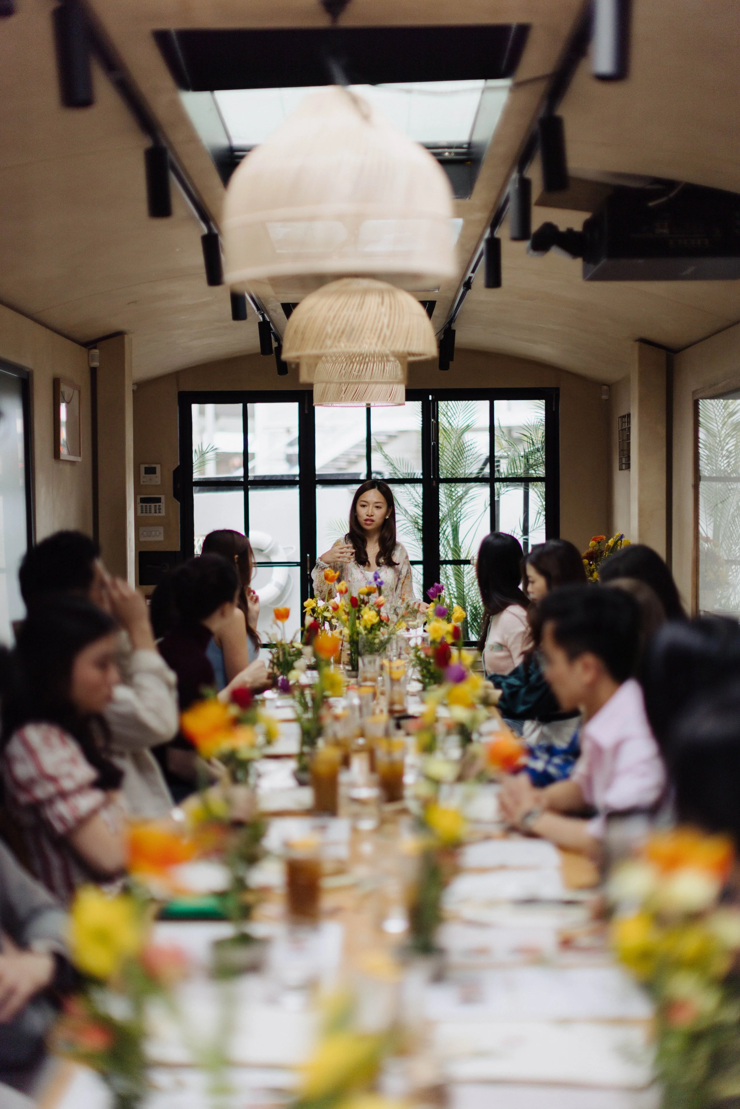 A woman standing at the head of a long dining table with colorful flowers, with people seated on both sides, in a room with large windows, pendant lights, and a natural decor style.