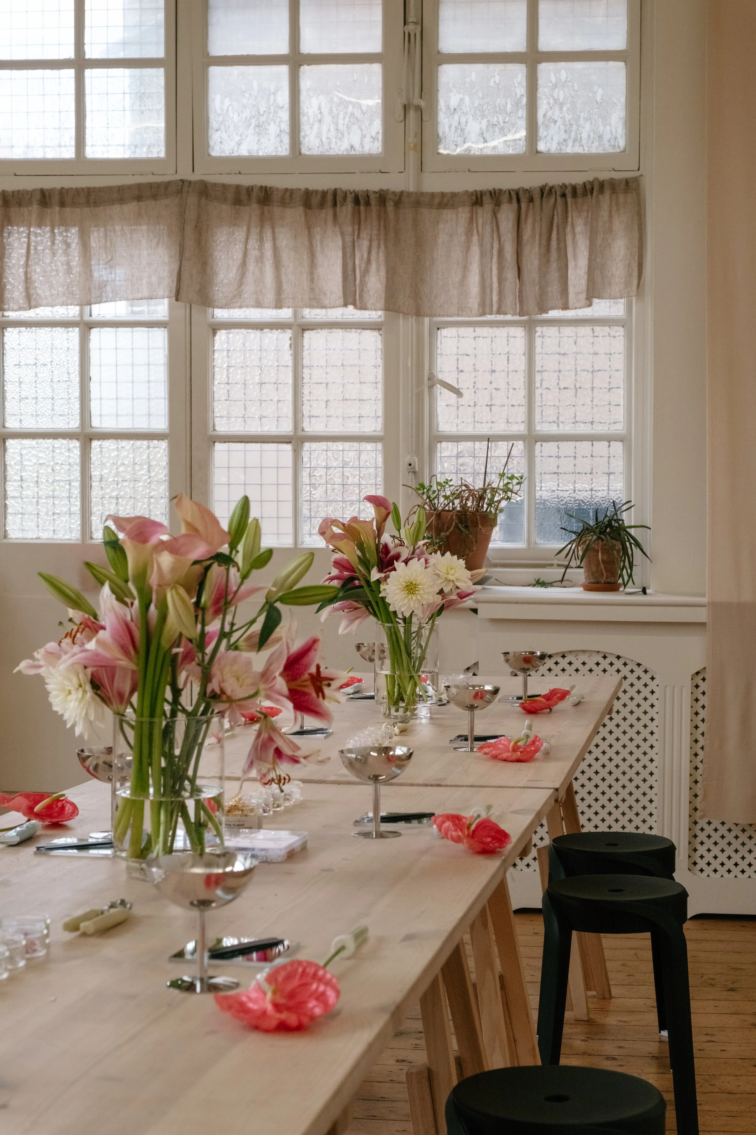 A decorated dining table set for a celebration in front of a large window with curtains. The table has floral centerpieces with pink and white flowers, and tableware including glasses, candles, and floral-shaped candle holders.