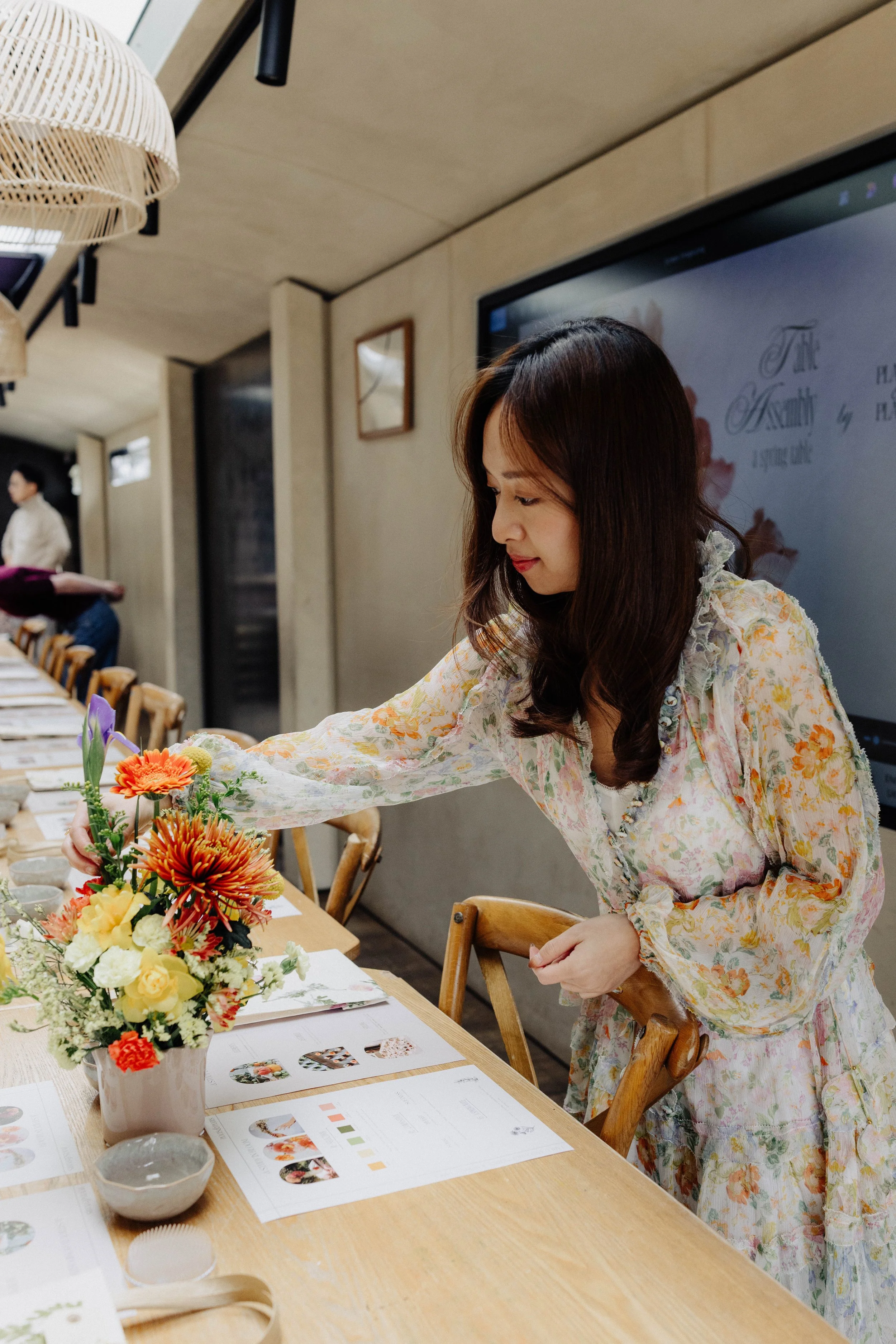 A woman with long dark hair in a floral dress arranging flowers at a table decorated with various colorful flower arrangements and papers.