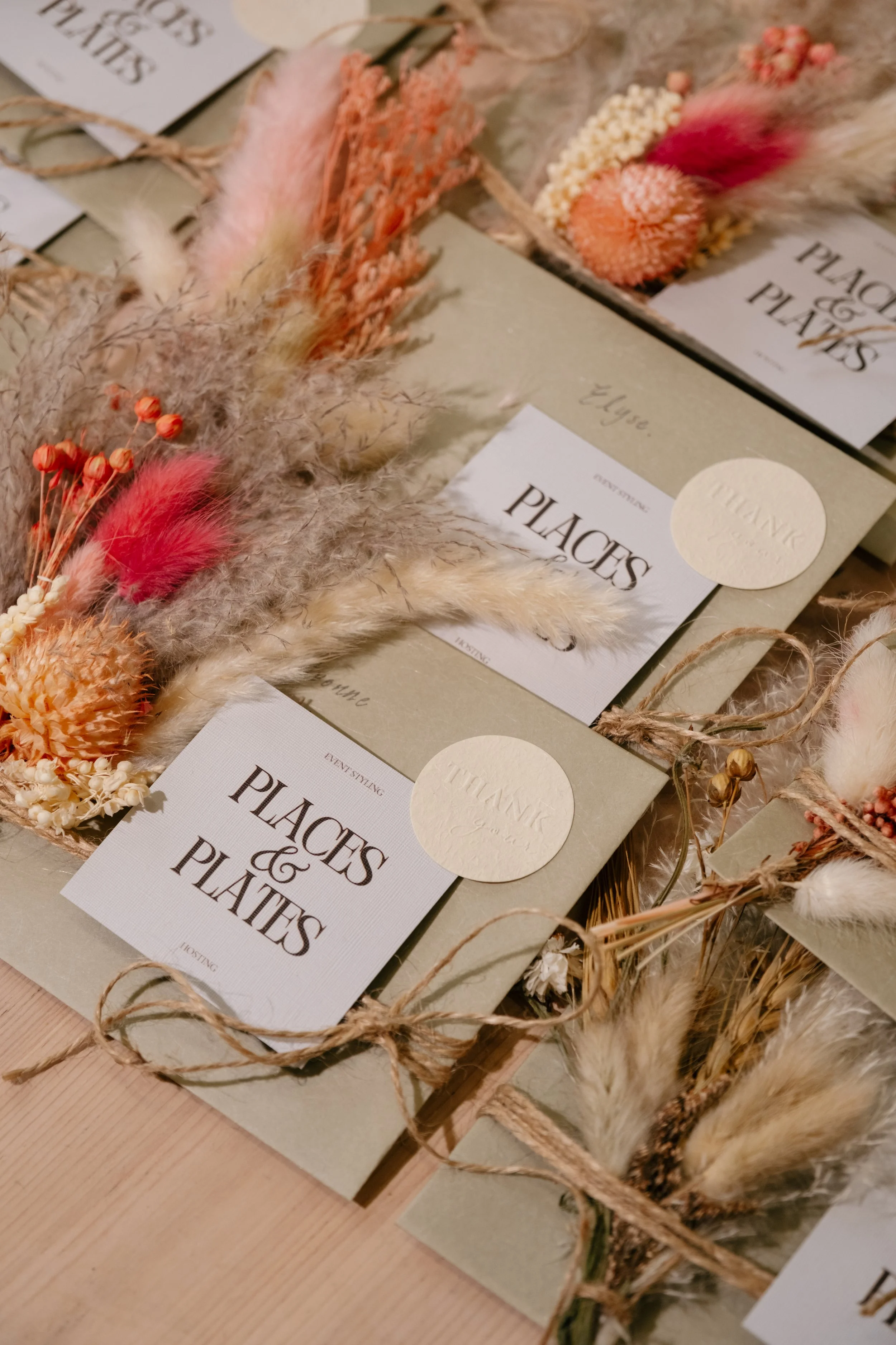 Wrapped packages with labels reading ''Placés & Plates'' decorated with dried flowers and grasses in soft pink, cream, and orange tones.