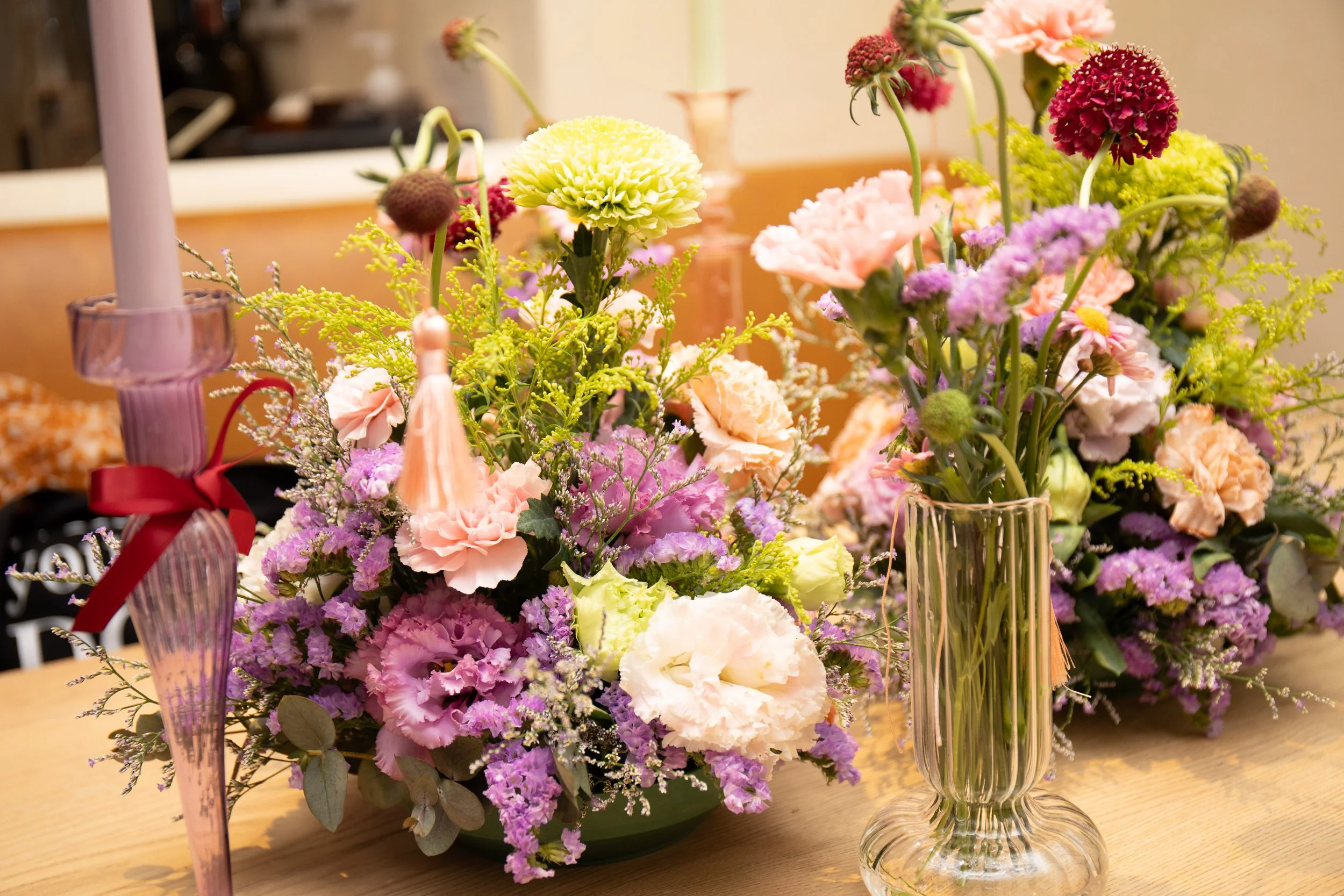 Colorful flower arrangement with pink, purple, yellow, and white flowers on a wooden table, with vases and candle holders in the background.