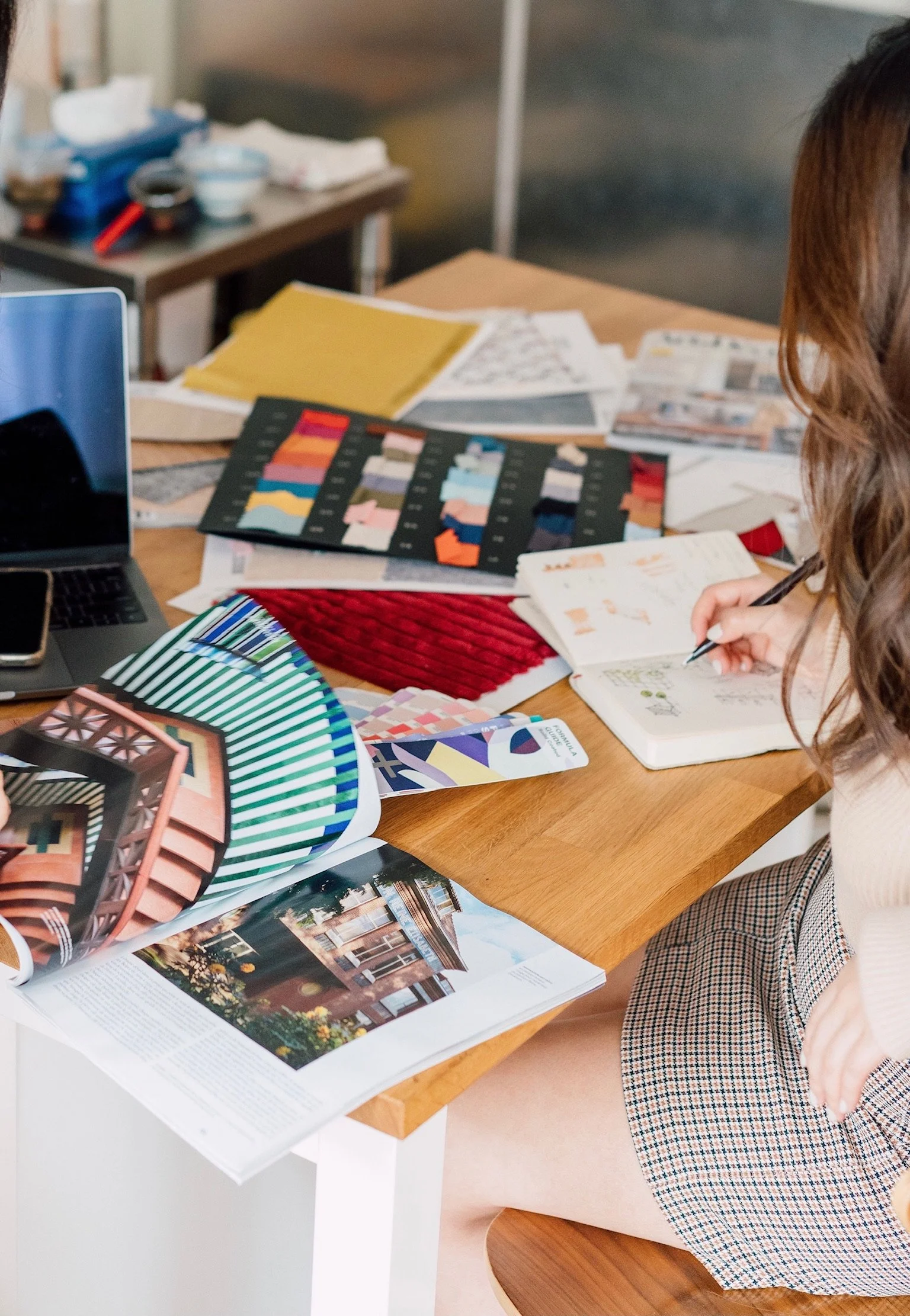 A person sitting at a desk reviewing an open magazine with interior design images, surrounded by color swatches, sample fabrics, and printed material, with a laptop in the foreground and open notebooks or papers on the desk.