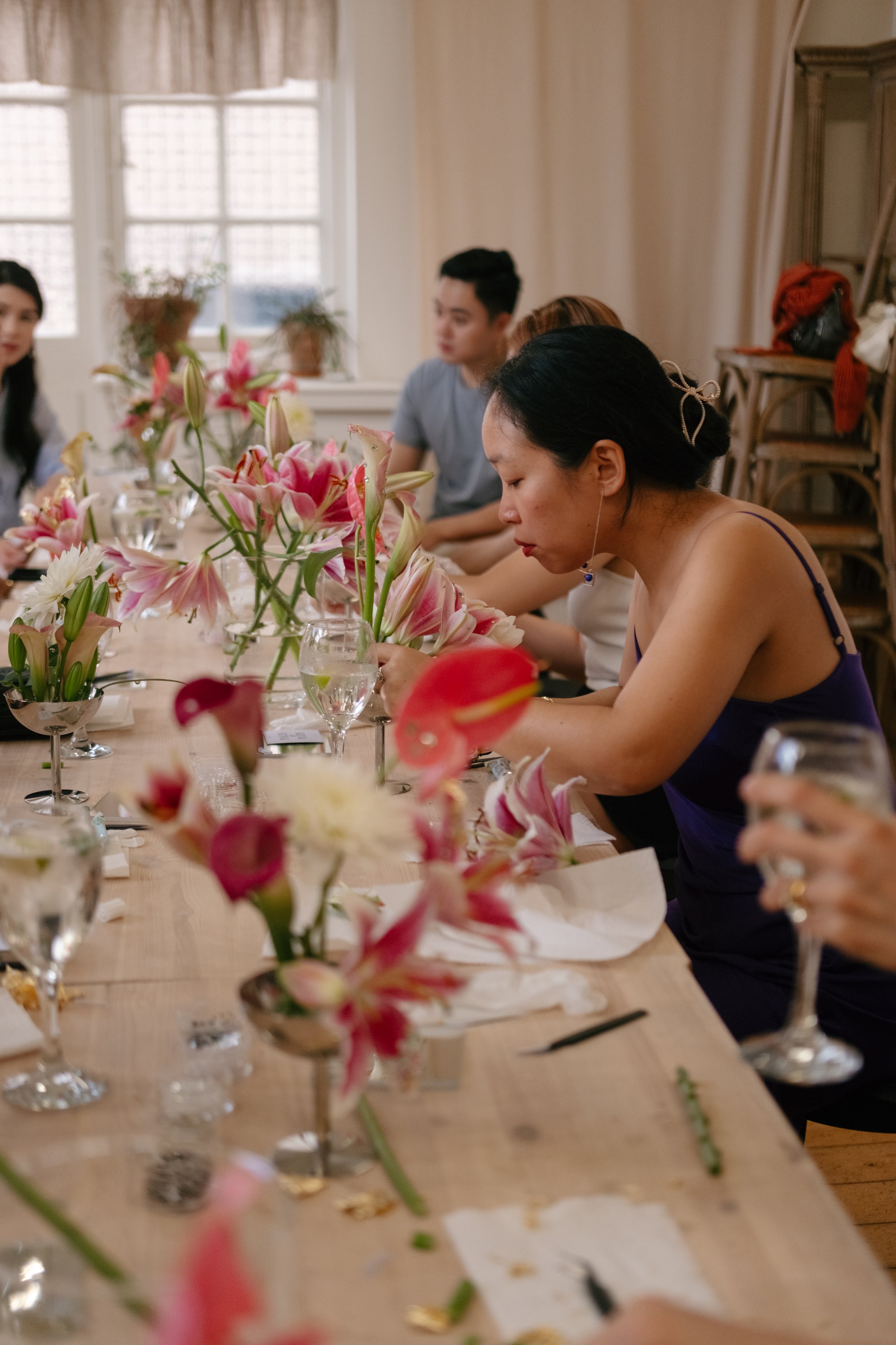 People sitting at a table decorated with pink and white lilies and glasses of water for a gathering or meal.