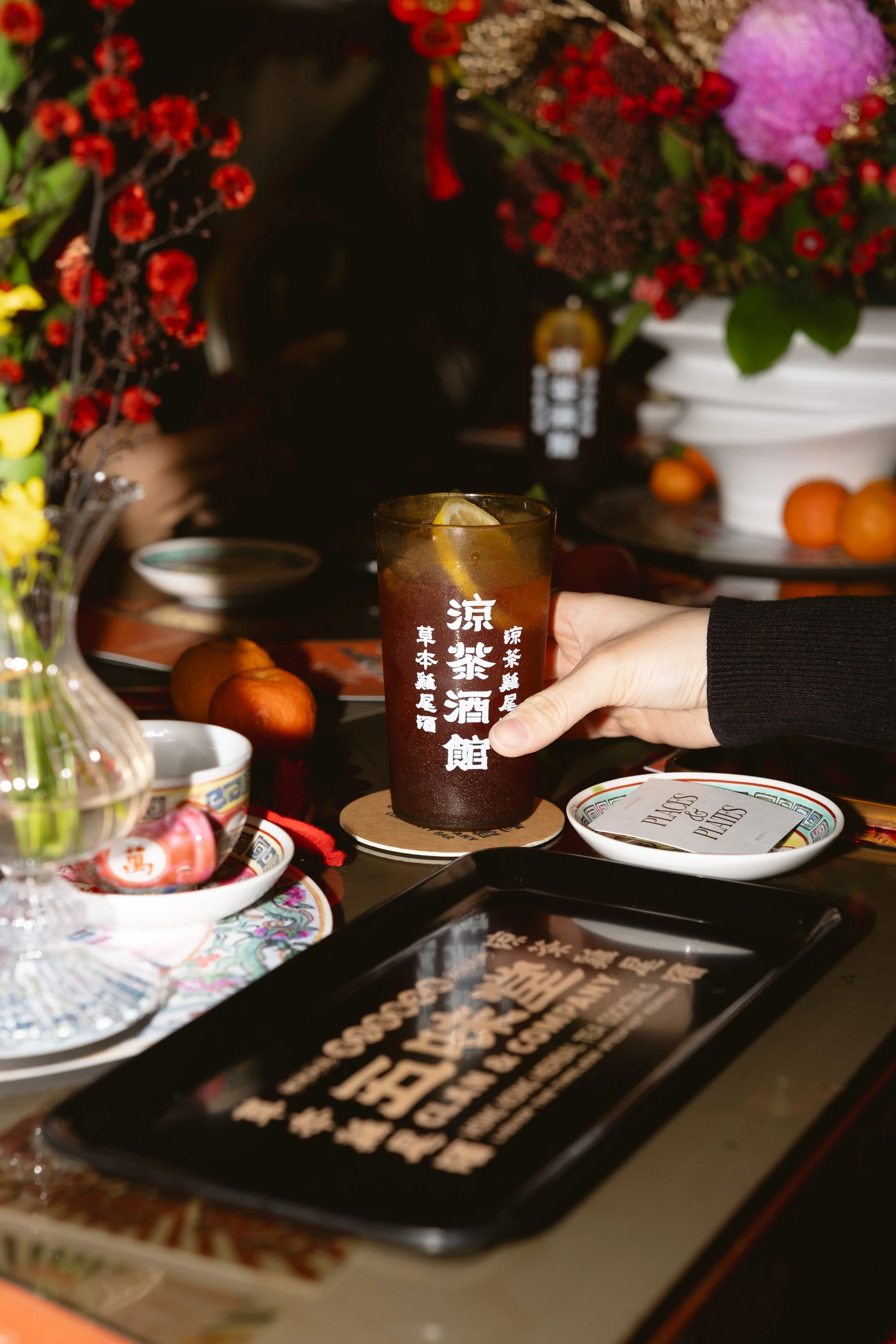 A hand holding a tall glass of iced tea with lemon slices on a decorated table with flowers, fruit, and various dishes.