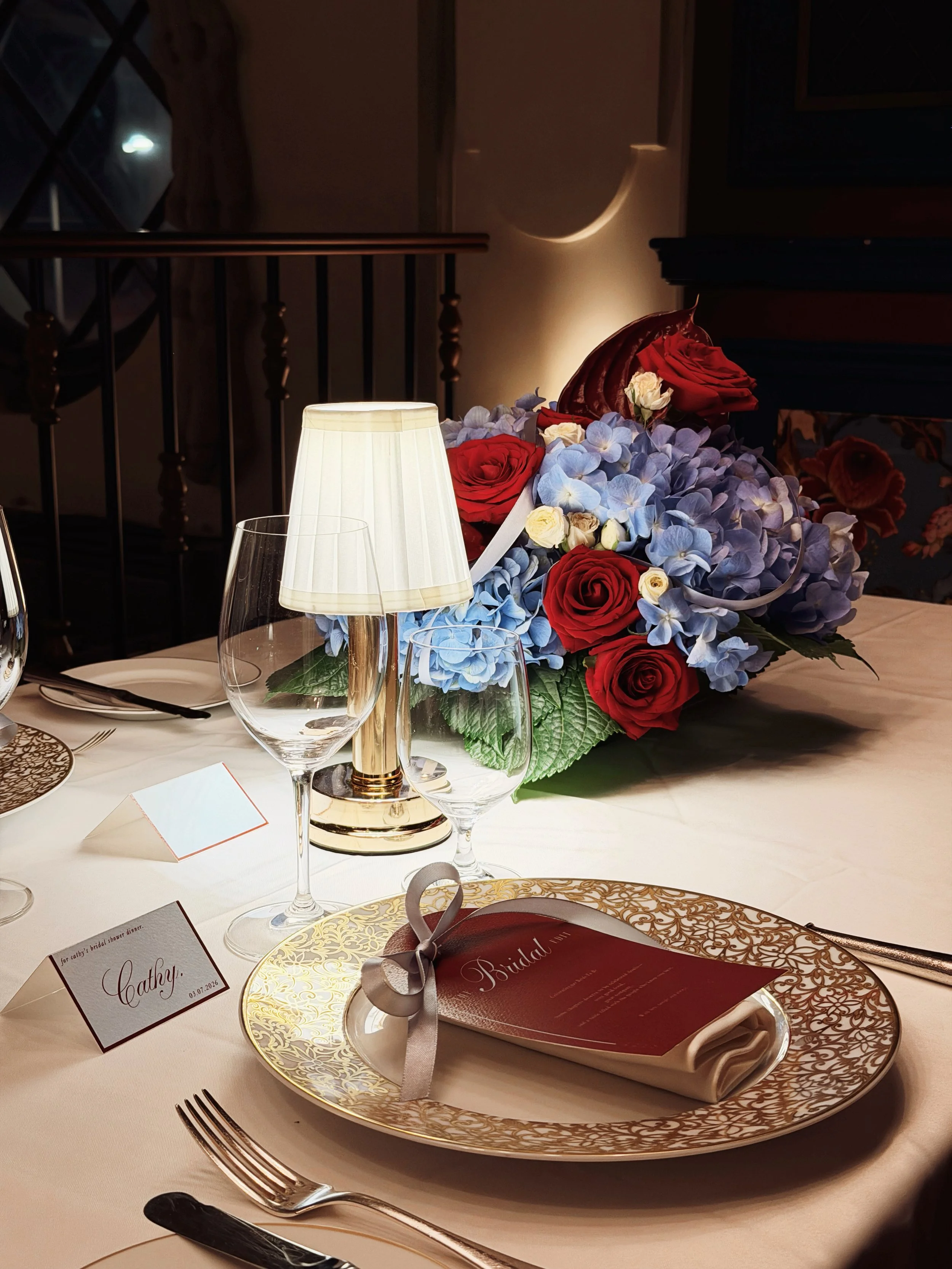 A formal table setting with a bouquet of red and blue flowers, a small lamp, wine glasses, a plate with a tied napkin and a menu, and a name card with the name 'Cathy' for a bridal shower dinner.