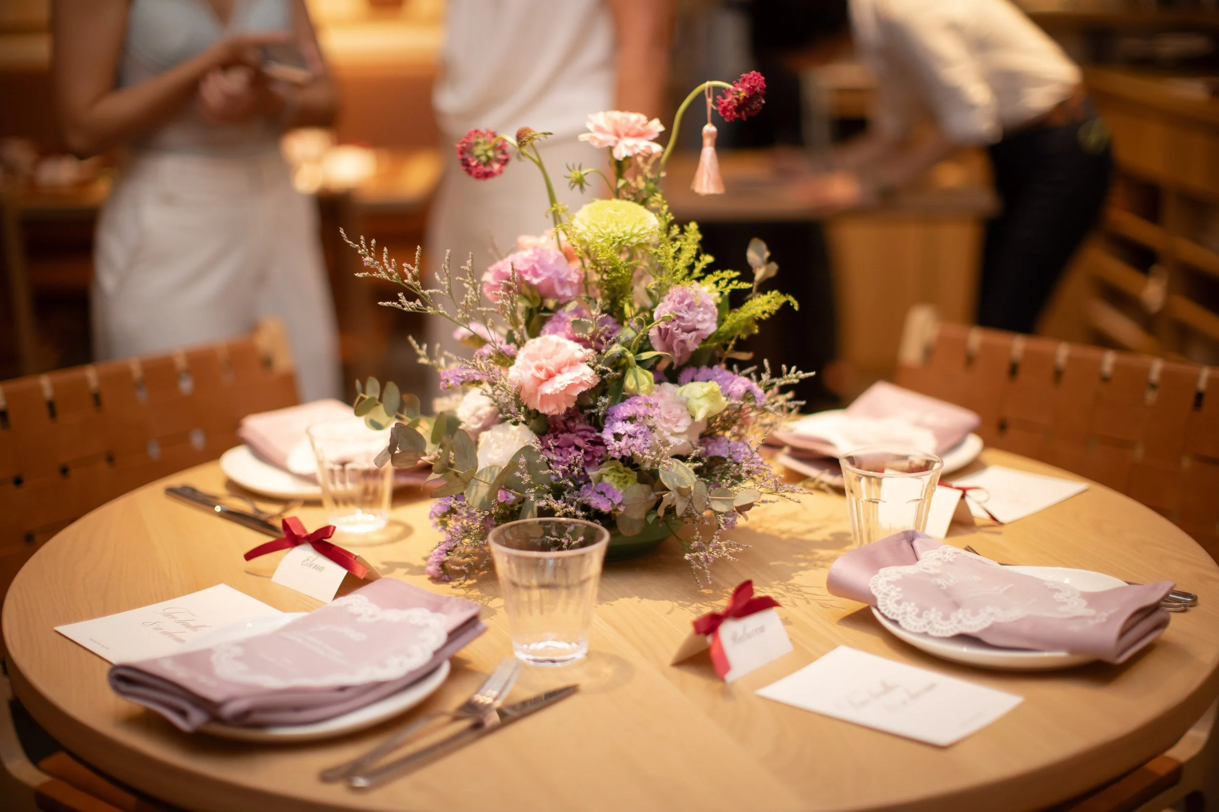 A floral centerpiece with pink, purple, and green flowers on a round wooden table set for a meal, with place cards, napkins, and glasses. In the background, people are standing and using their phones in a warmly lit setting.