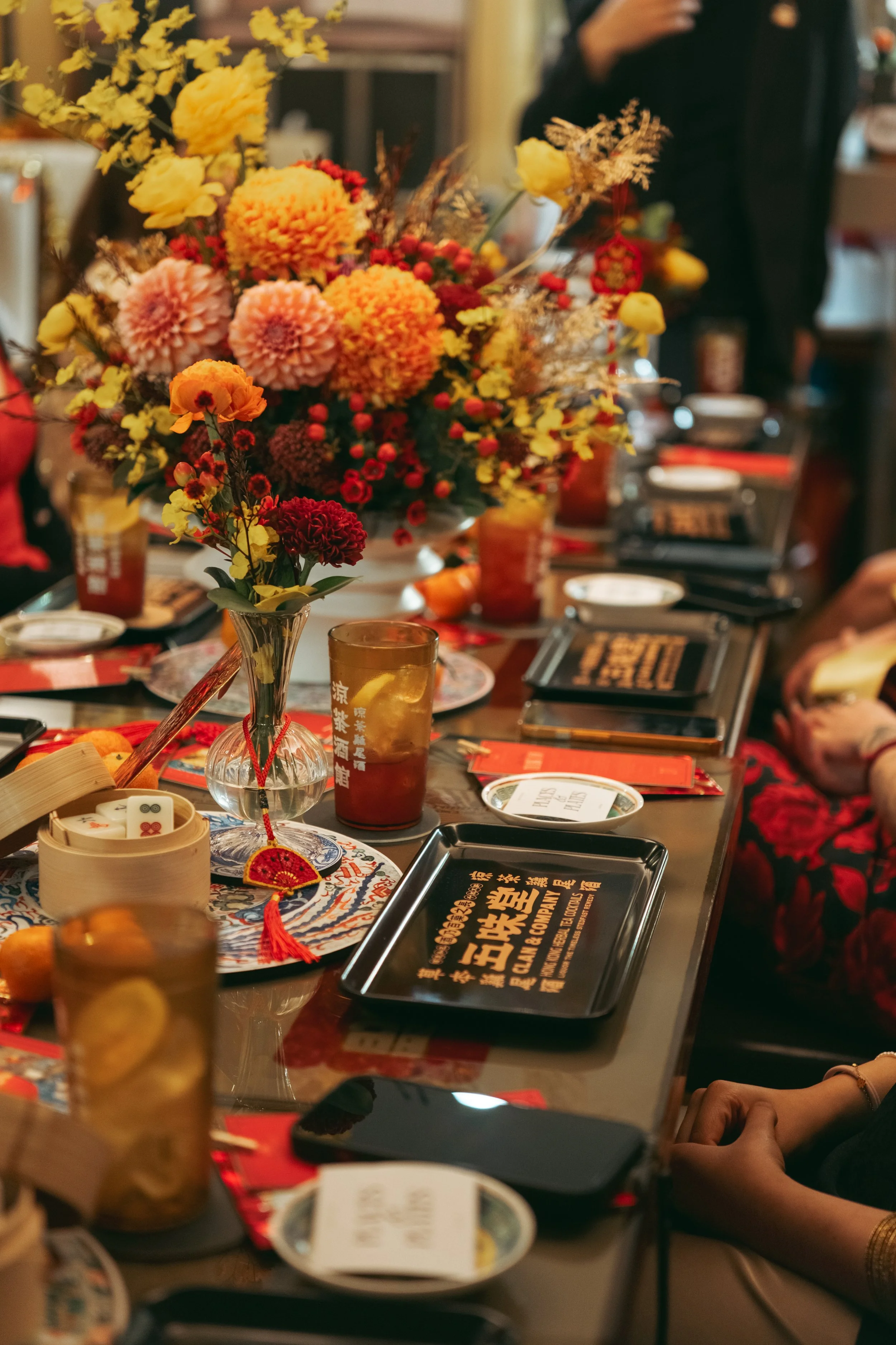 A table decorated with a large colorful flower arrangement featuring yellow, orange, pink, and red flowers. The table also has several drinks, black containers with Chinese characters, small plates, and a decorative item with a red tassel. There are people in the background, some with hands visible, indicating a social gathering or celebration.