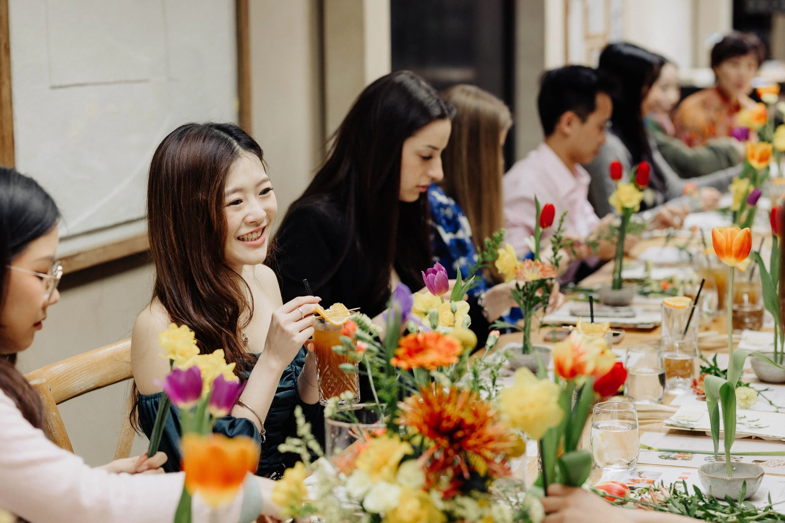 People sitting at a long table with floral centerpieces, enjoying a meal together in an indoor setting.