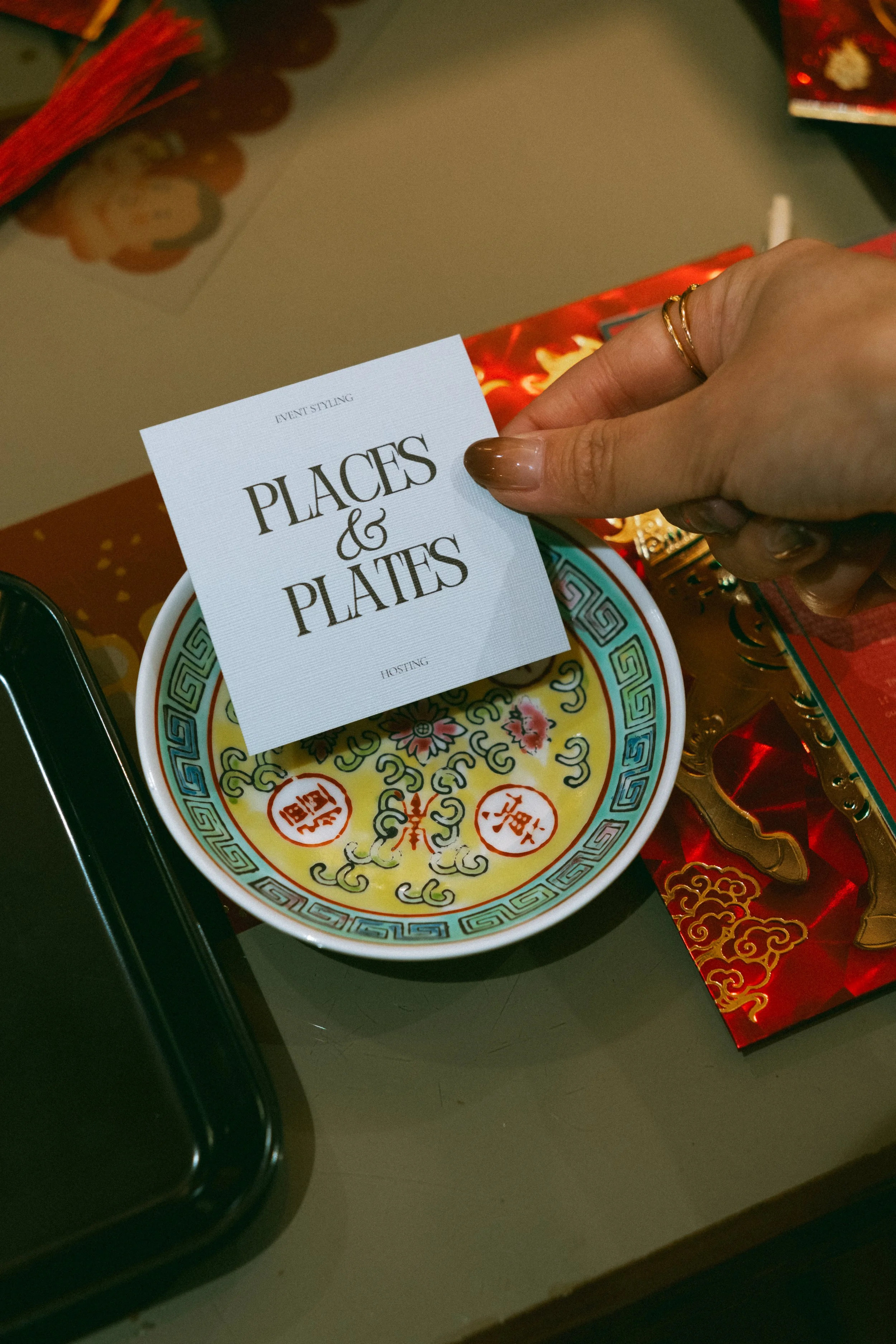 A hand holding a white card with black text that reads 'PLACES & PLATES' above a decorative ceramic plate with Chinese characters and intricate patterns, surrounded by red and gold decorations.