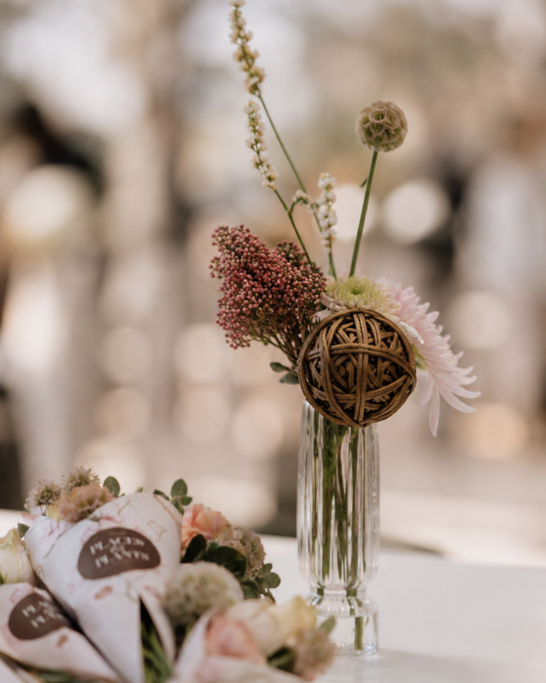 A glass vase with a small arrangement of pink and white flowers, including a spherical decorative element made of woven wood, with a blurred background.