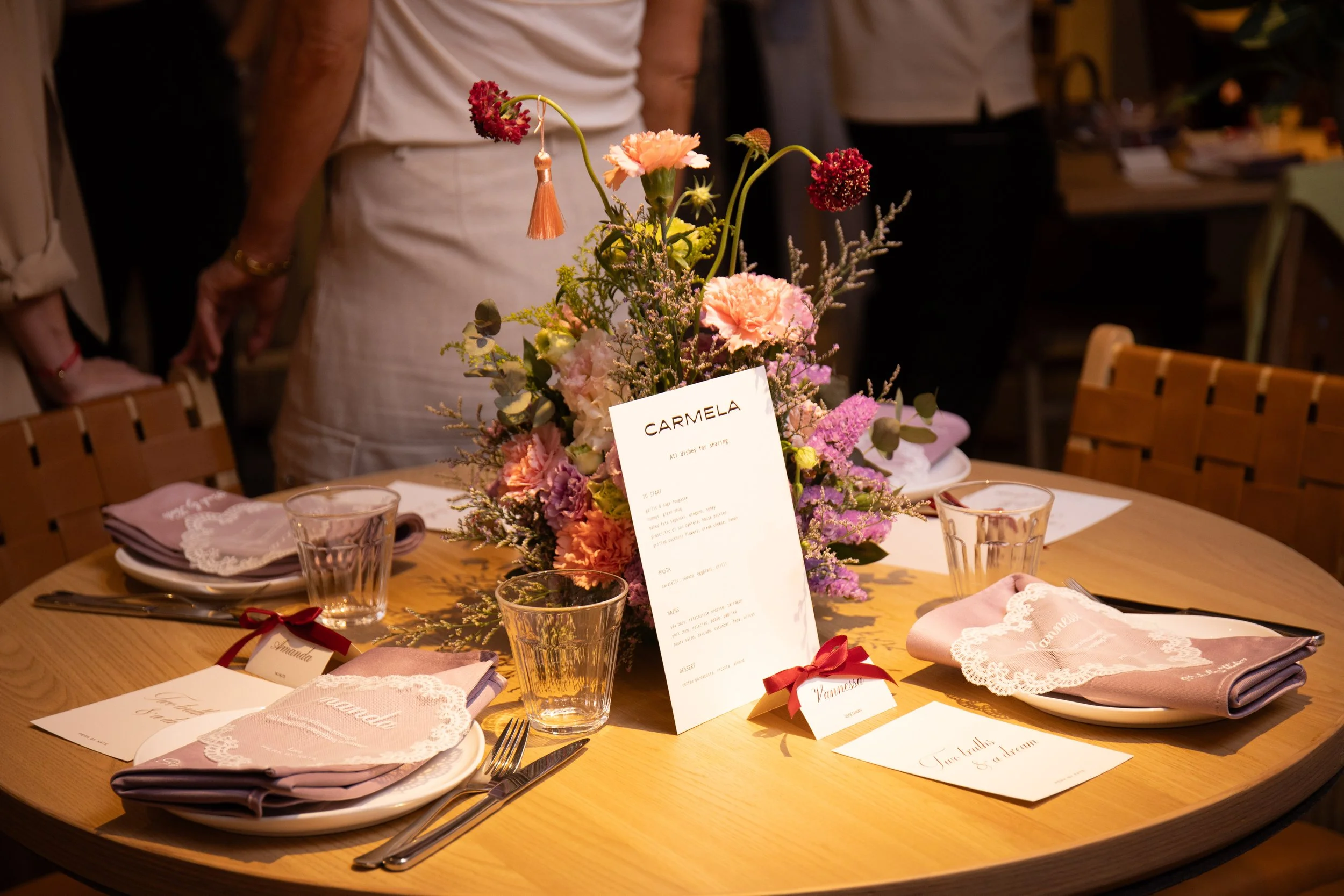 A table decorated with a pink and purple flower arrangement, place cards, and pink napkins with lace borders. The table settings include clear glasses and cutlery. In the background, people are standing, and there is a menu card in the center of the table with the name 'Carmela' on it.
