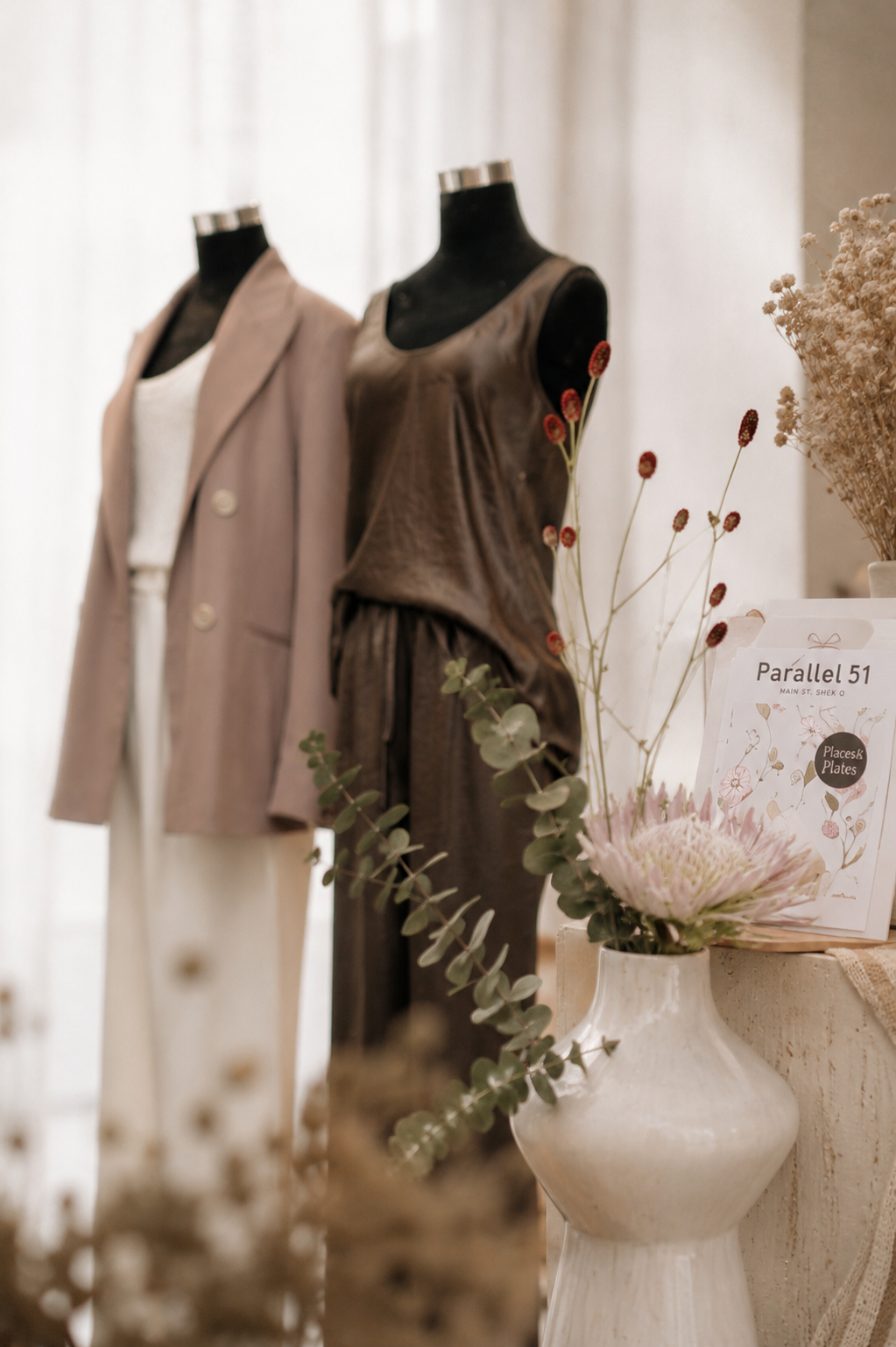 Two mannequins dressed in neutral-colored outfits, one in a beige blazer and white dress, the other in a brown sleeveless top and matching pants. A bouquet of flowers and greenery is also visible in the foreground.
