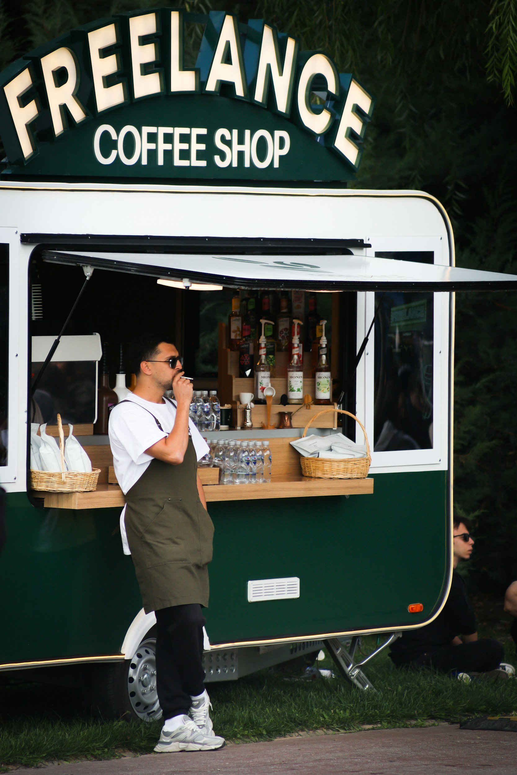 A man wearing sunglasses and a white shirt with an apron is standing outside a green trailer with a coffee stand, drinking from a straw. The trailer has a sign that says 'FREELANCE COFFEE SHOP' on top.