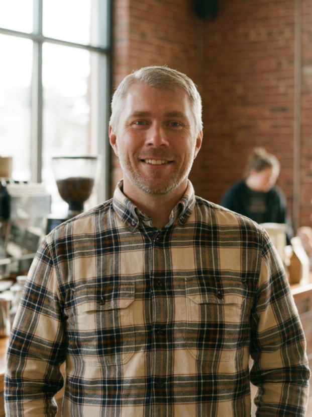 A smiling middle-aged man with gray hair and beard wearing a plaid shirt inside a coffee shop.